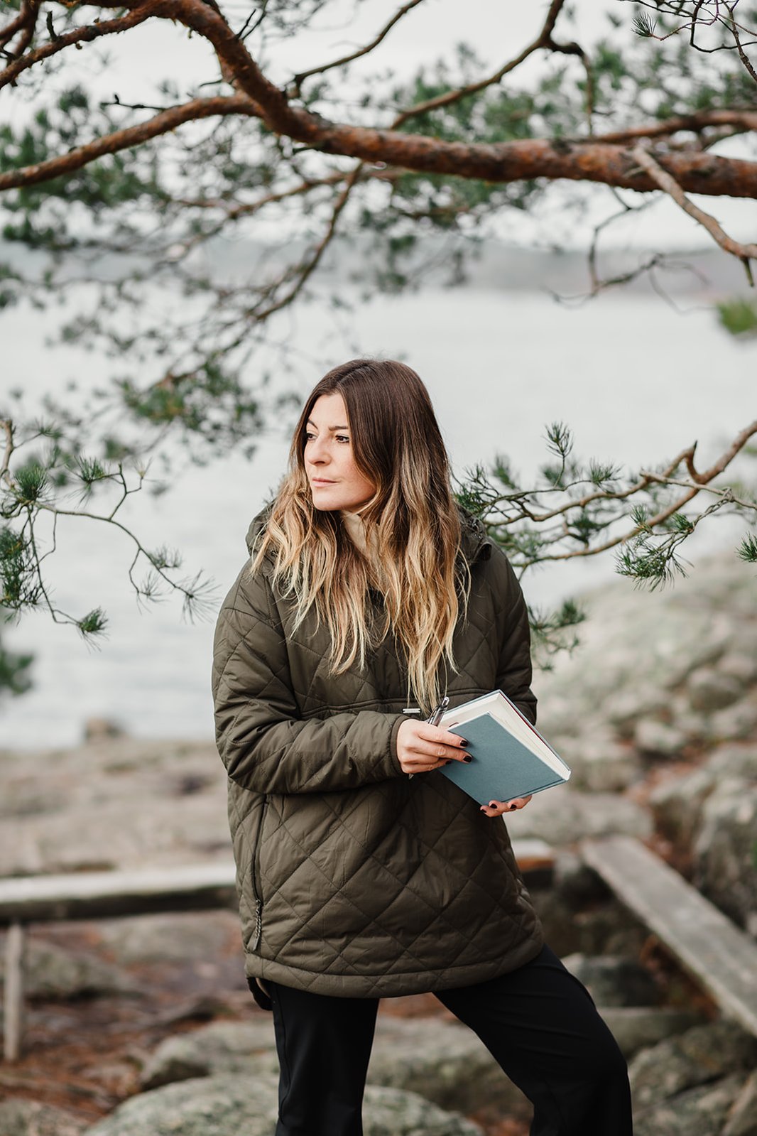 Kvinna med långt hår, grön quiltad jacka under ett träd i skärgården med en blå anteckningsbok och penna i handen, tittar till vänster i bilden. Bakom vatten och en träbänk.