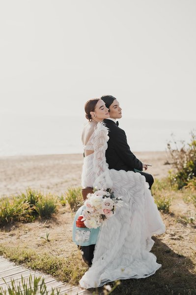 Par sitter tätt tillsamman på en vespa nära en strand. Hon i vit lång klänning och brudbukett i handen. Han fram i svart kostym.
