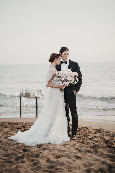 Brudpar står på sandstrand, bakom ett bord och vattnet vid havet. Hon klädd i vit lång klänning och blommor i handen, han i svart kostym och vit skjorta.