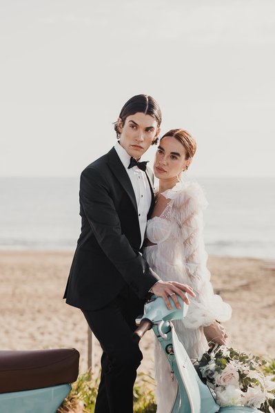 Par står på en strand framför en ljusblå vespa. Han i mörkt hår och mittbena klädd i mörk kostym, hon i vit lång klänning och blommor i handen.
