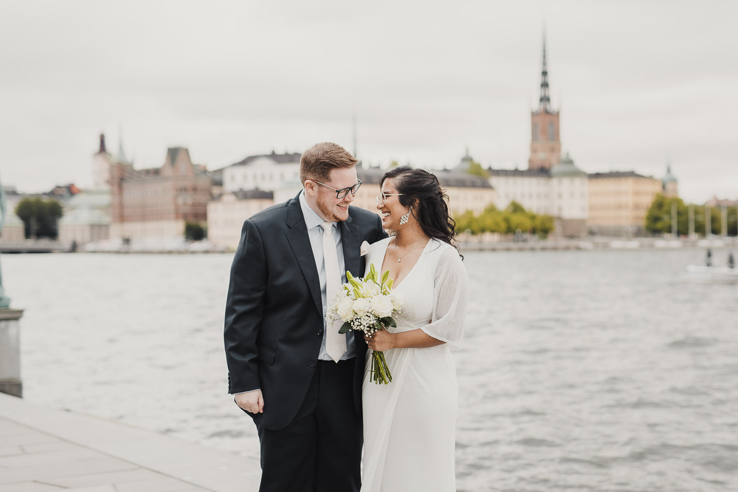 Man i blå kostym och glasögon står tätt tillsammans och pratar med en kvinna i vit klänning, mörkt hår och glasögon. Bakom dem vattnet från Mälaren och husfasaderna på ön Riddarholmen.