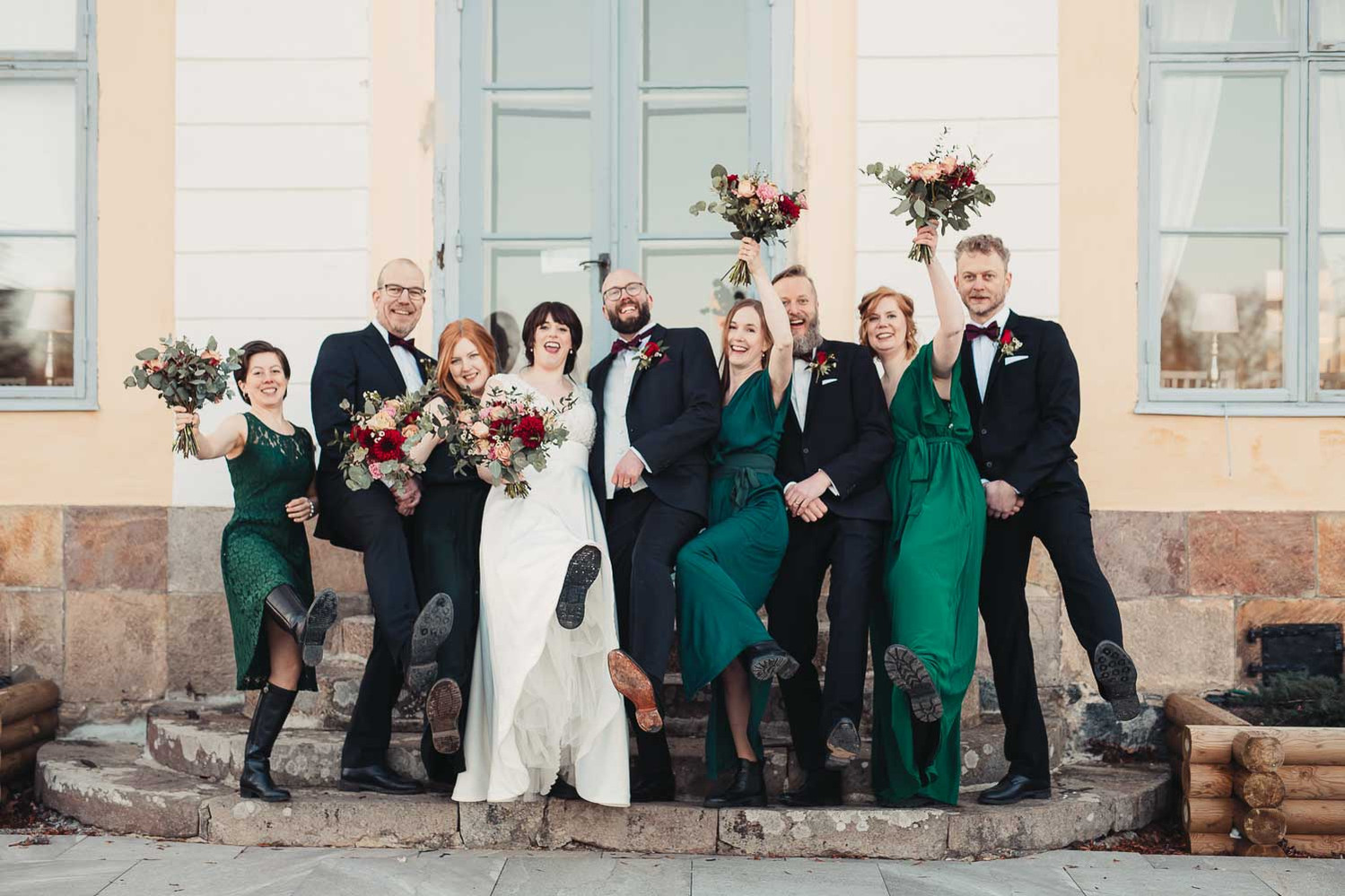 Brudpar och brudfölje står på en trappa utanför en herrgård. Brudtärnor  i gröna klänningar och håller varsin bukett blommor i handen och männen i mörk kostym. Alla sträcker upp ett ben och en hand i luften.