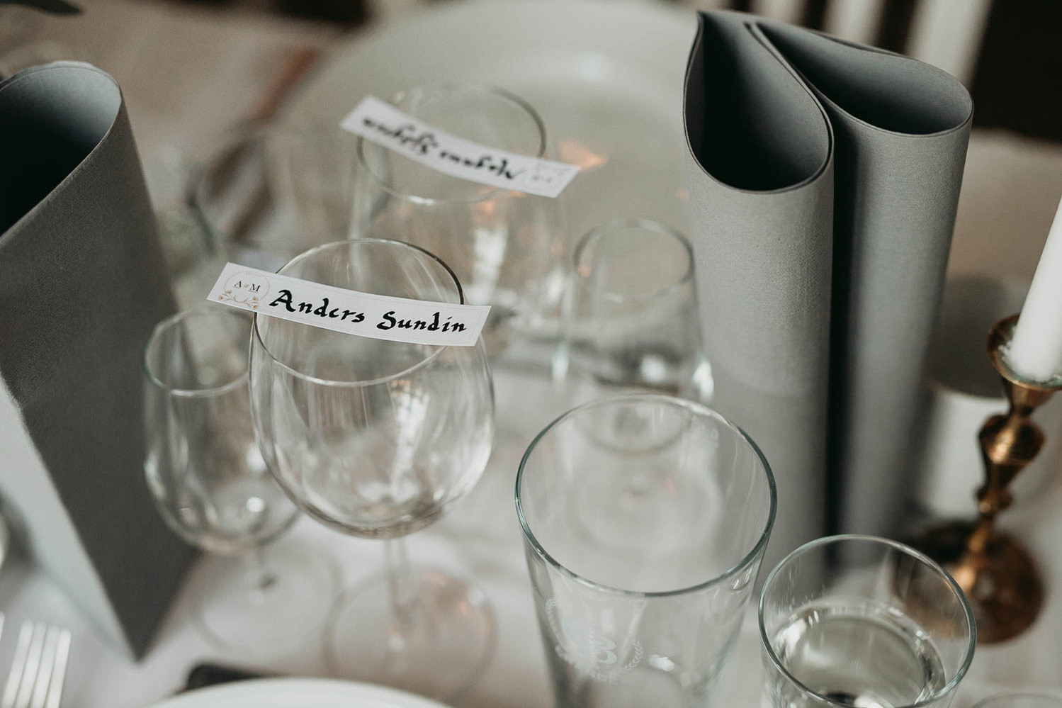 Vinglas på dukat bord med vit duk. Glas för nubbe, vattenglas, grå vilta servetter. Vita skylt med namnet Anders  S i svart text.