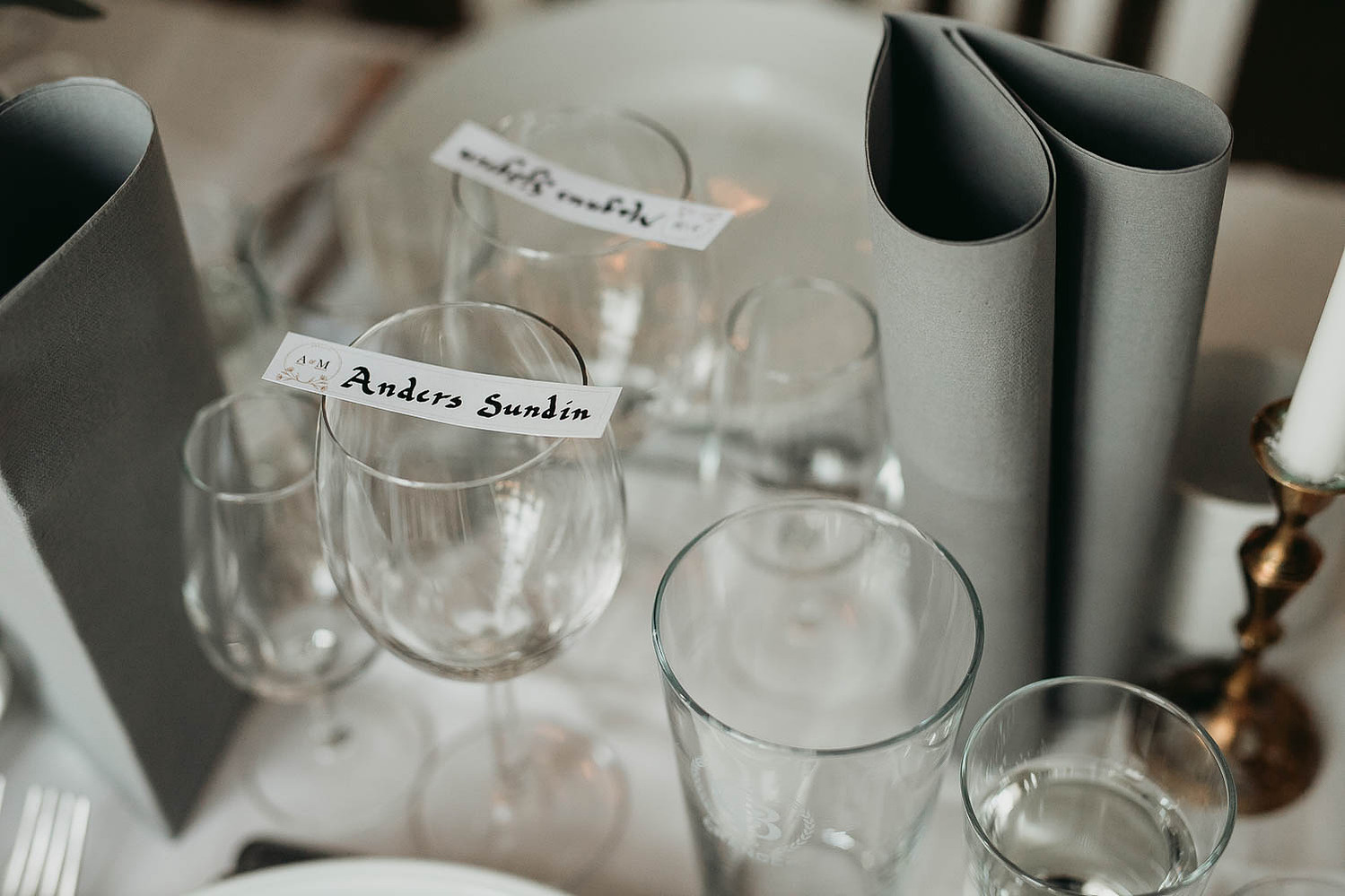 Vinglas på dukat bord med vit duk. Glas för nubbe, vattenglas, grå vilta servetter. Vita skylt med namnet Anders  S i svart text.