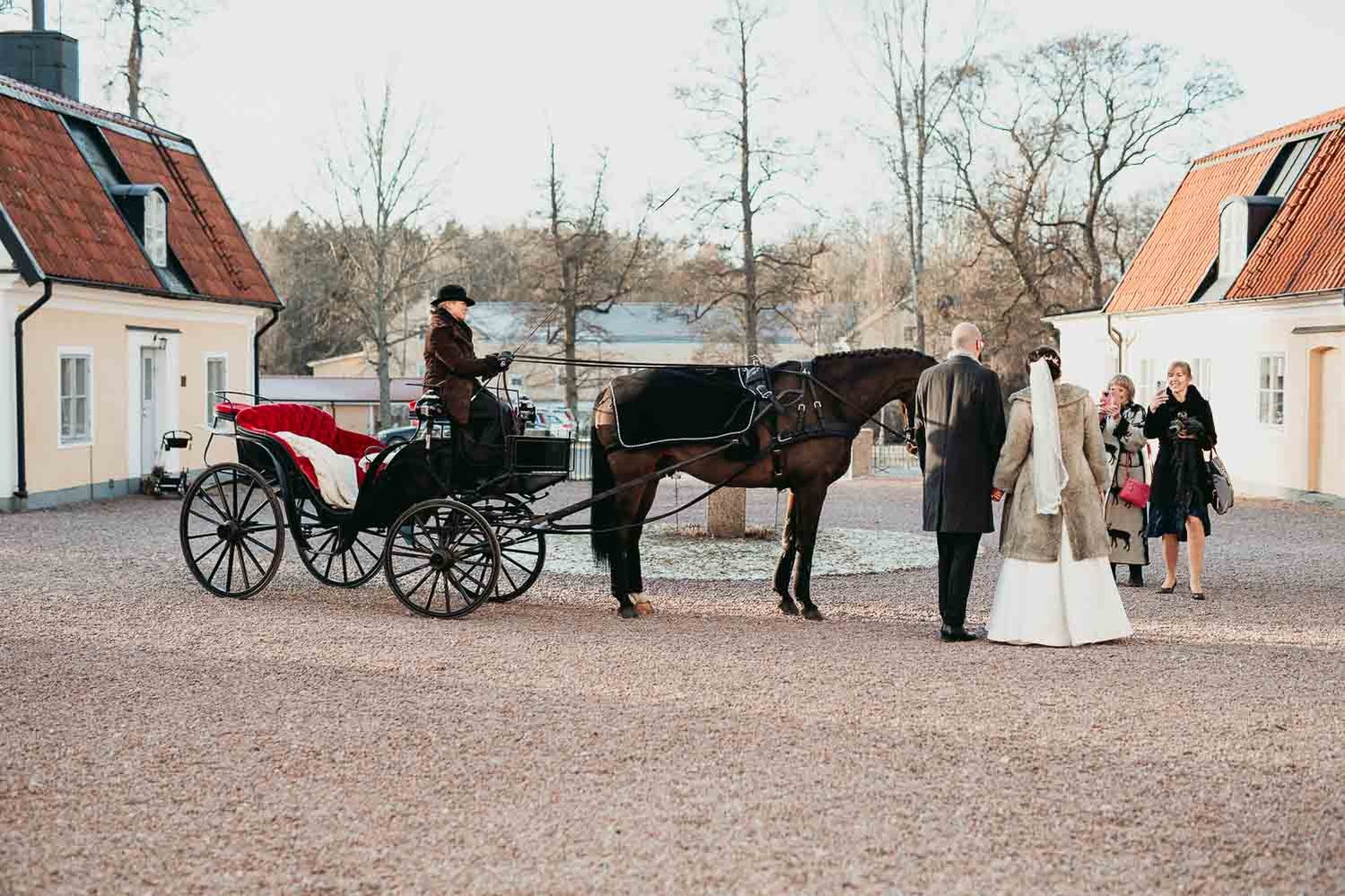 Brudpar står på en gårdsplan bredvid en häst med vagn och kusk medan några gäster tar foton med mobilen.