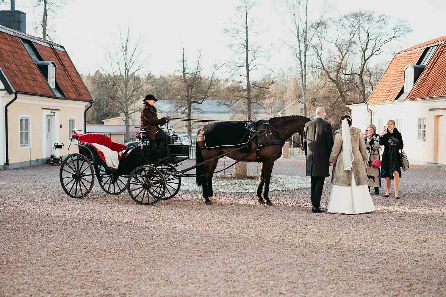 Brudpar står på en gårdsplan bredvid en häst med vagn och kusk medan några gäster tar foton med mobilen.