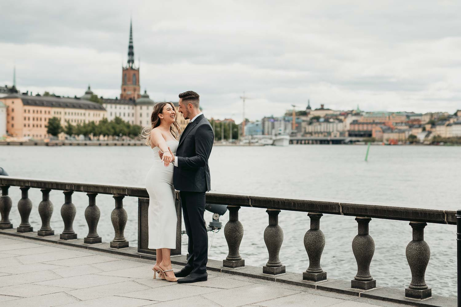 Par håller om varandra utomhus i stan. Hon i vit klänning och han i mörk kostym. Bakom dem svart räcke och vattnet i Mälaren och husfasader från Riddarholmen.