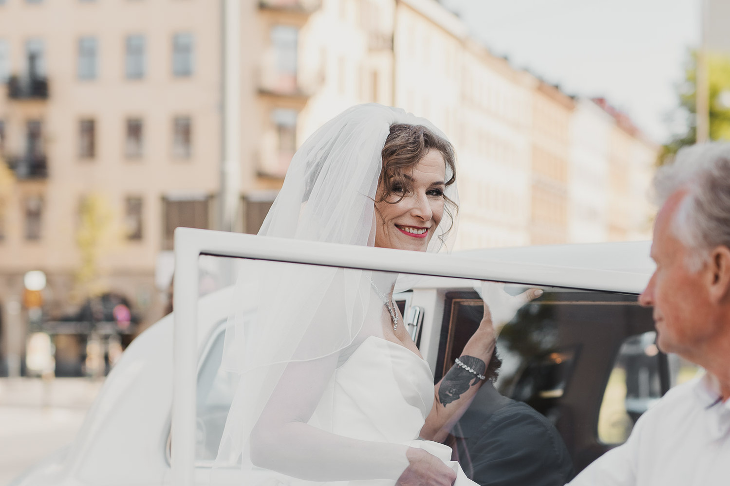 Brud i vit klänning och brudslöja i håret kliver i en vit limousine, chauffören håller upp dörren, i bakgrunden gult höghus i stan. 