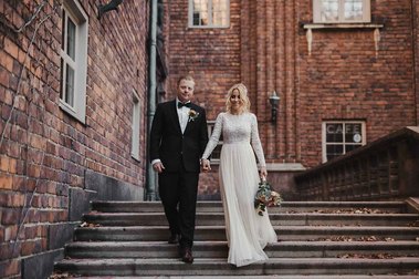 Bröllopspar går ner för en trappa med höstlöv i bakgrunden röd tegelfasad med fönster. Han i mörk kostym, hon i vit klänning och blommor i handen.