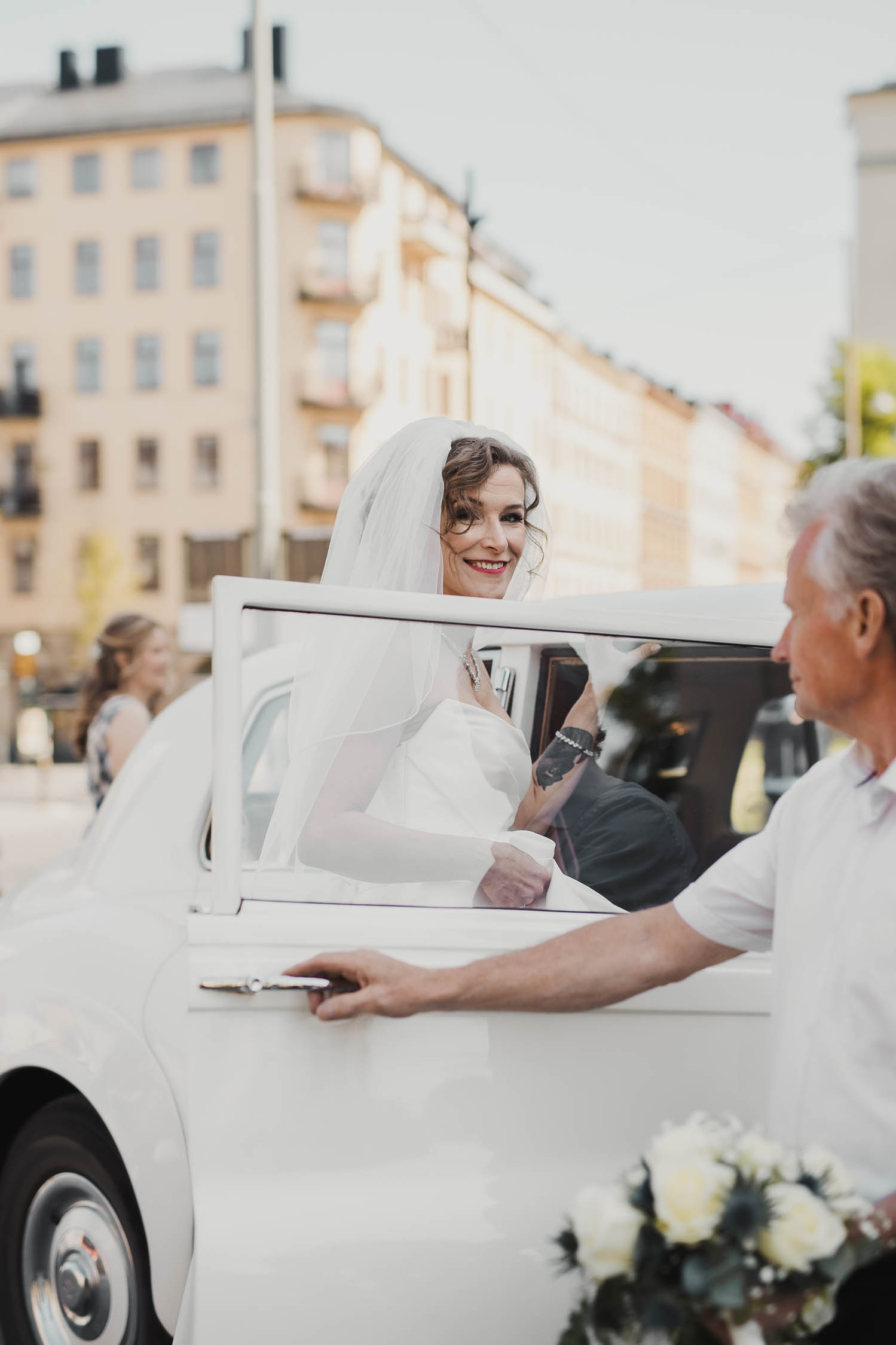 Brud i vit klänning och slöja kliver in vitt limousine, chauffören håller upp bildörren. Bakom gult höghus i stan.