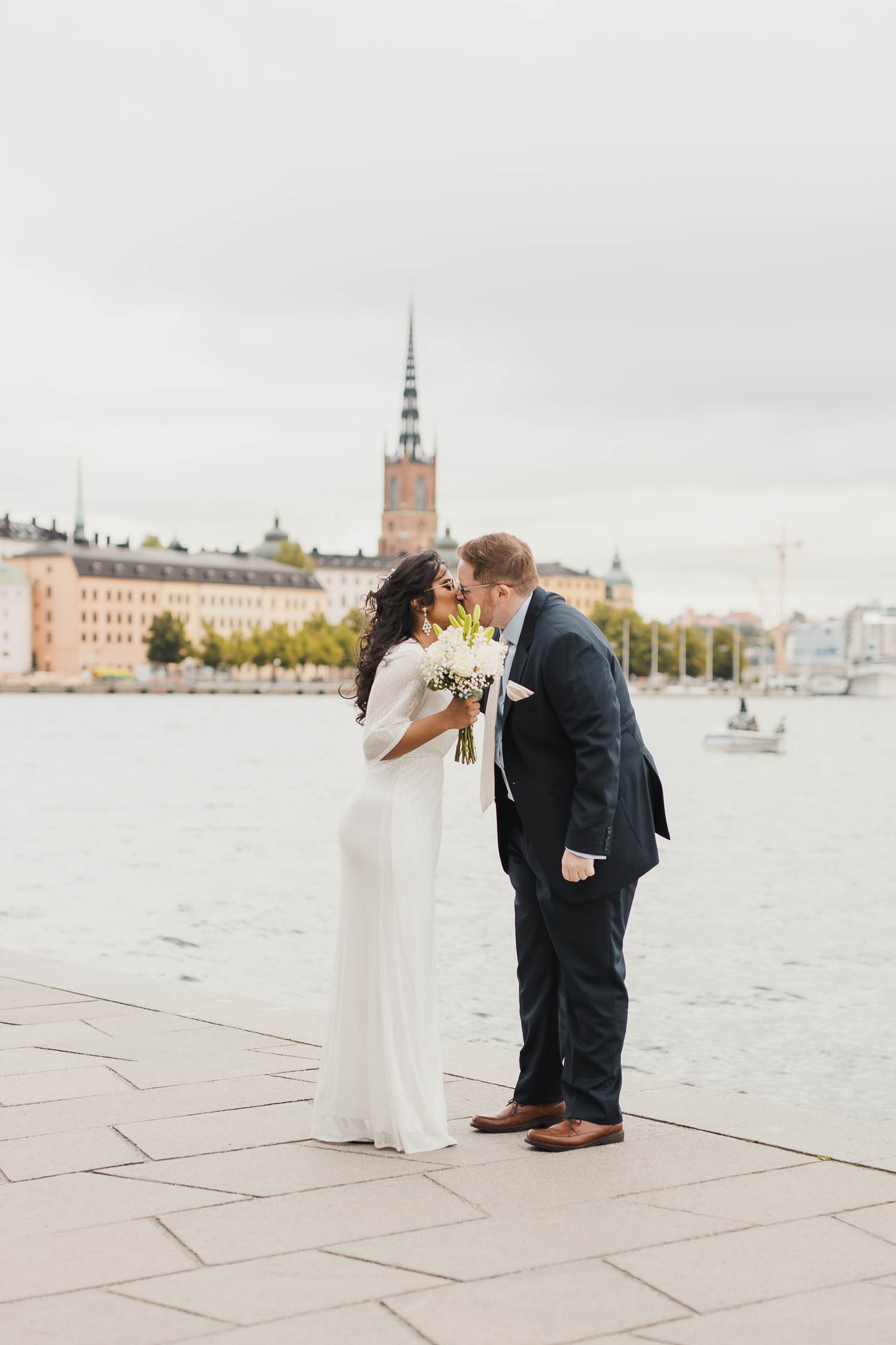 Man i blå kostym och slips kysser en kvinna i vit klänning och mörkt hår med brudbukett i handen. Dem står utomhus på gråa plattor vid kajen, bakom dem vatten och husfasader från ön mittemot.