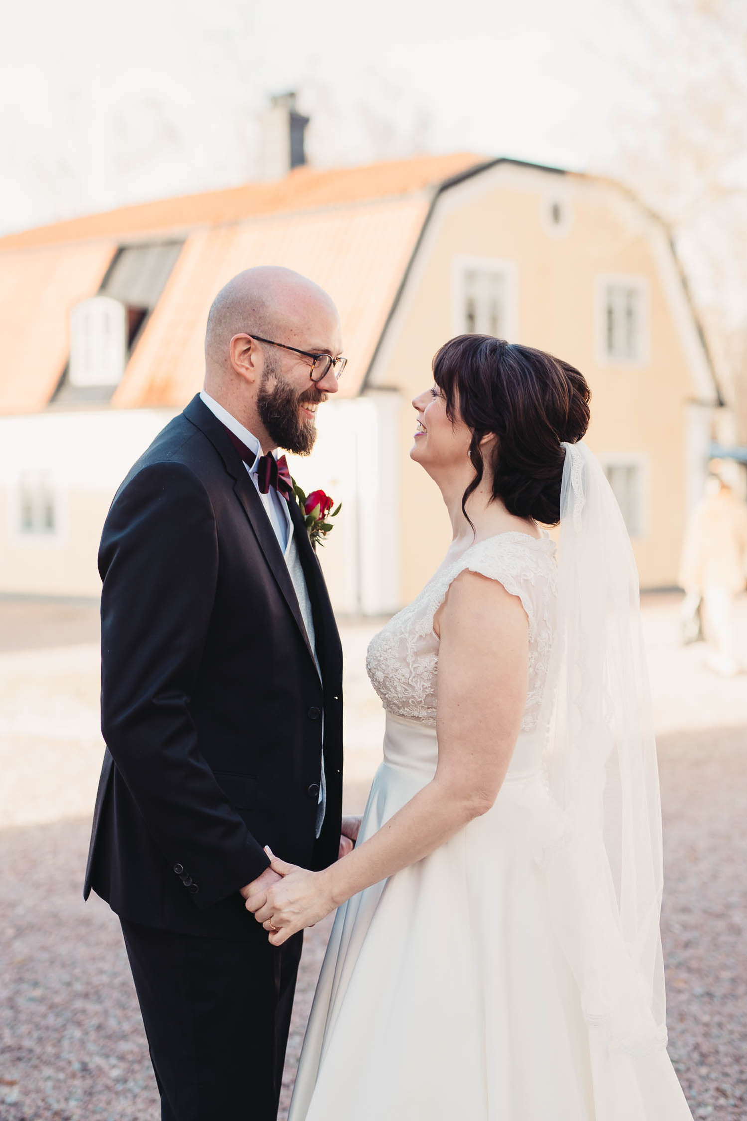 Man i mörk kostym, fluga och glasögon håller en kvinnas händer och ser henne i ögonen utomhus. Hon i mörkt uppsatt hår och slöja, vit lång  klänning utan ärm. Bakom dem hus med gul fasad och rött brutet tak.