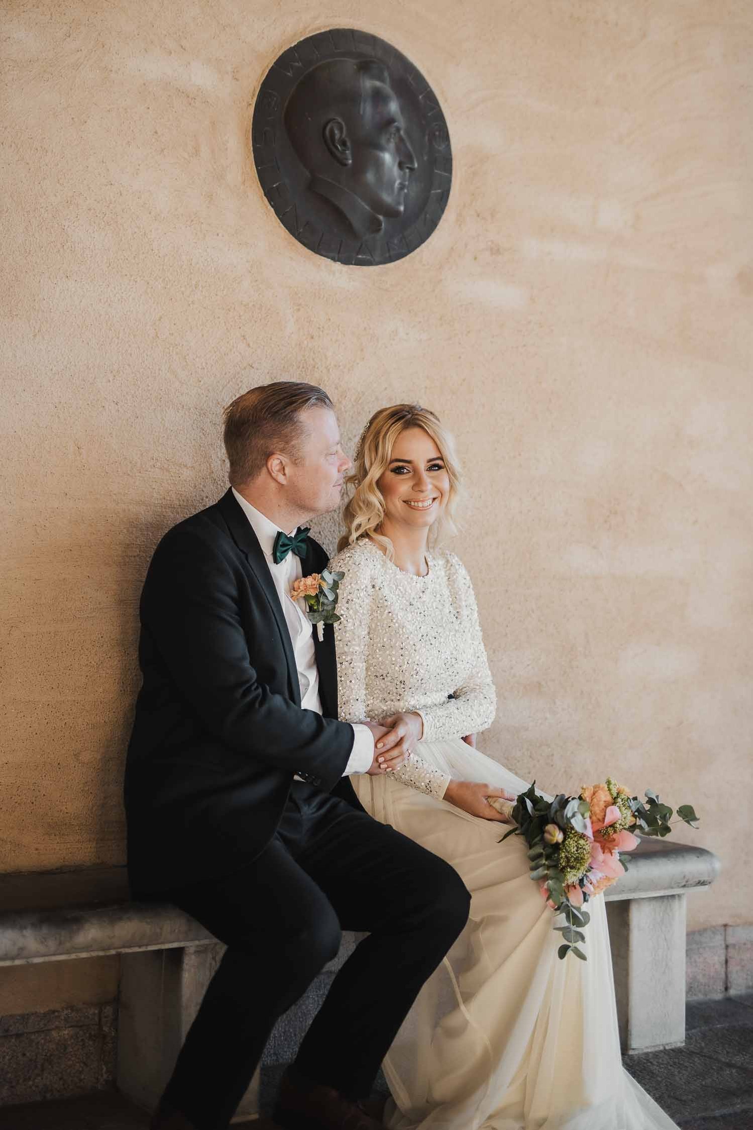 Bröllopspar sitter på en bänk, hon i vit lång klänning och brudbukett i handen. Han i mörkblå kostym och fluga. I bakgrunden sandstensfärgad vägg och en rund skulptur.