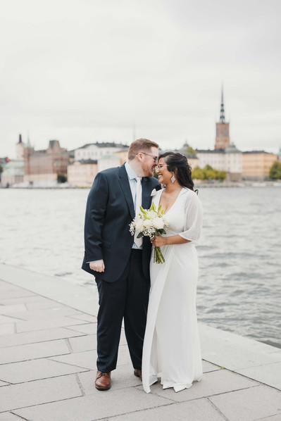 Brudgum säger viskar något i sin bruds öra, hon vit lång klänning och brudbukett, han i blå kostym. I bakgrunden vattnet och byggnader från Riddarholmen i Stockholm.