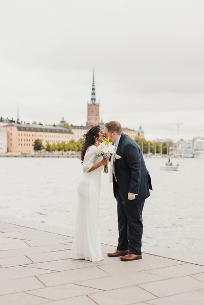 Brudpar kysser varandra utanför stadshuset, hon vit lång klänning och brudbukett, han i blå kostym. I bakgrunden vattnet och byggnader från Riddarholmen i Stockholm.