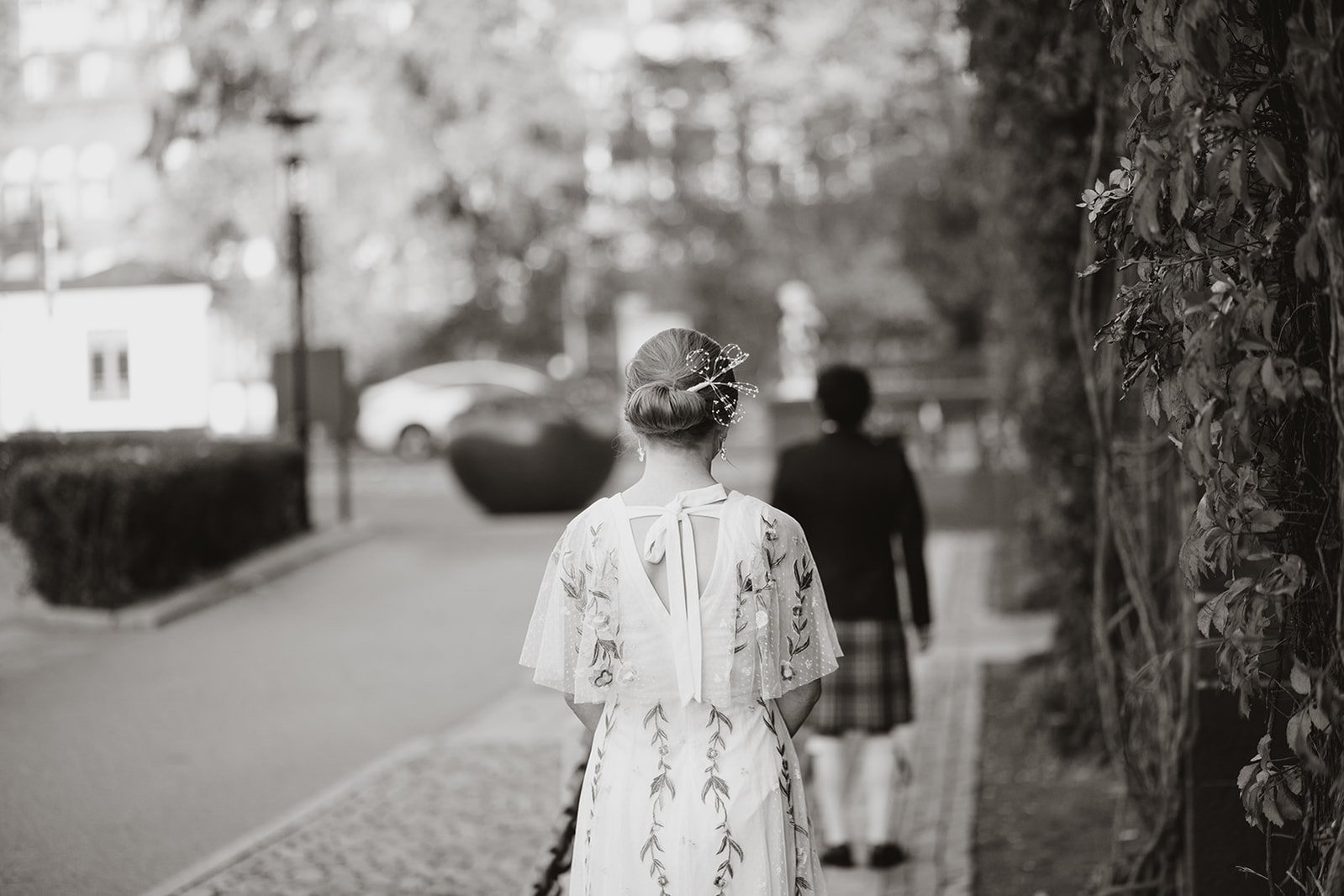 En kvinna med vit klänning och uppsatt hår står utomhus bakom en man med kavaj och kilt. På höger sida fasadens löv, på vänster sida väg och kullersten.