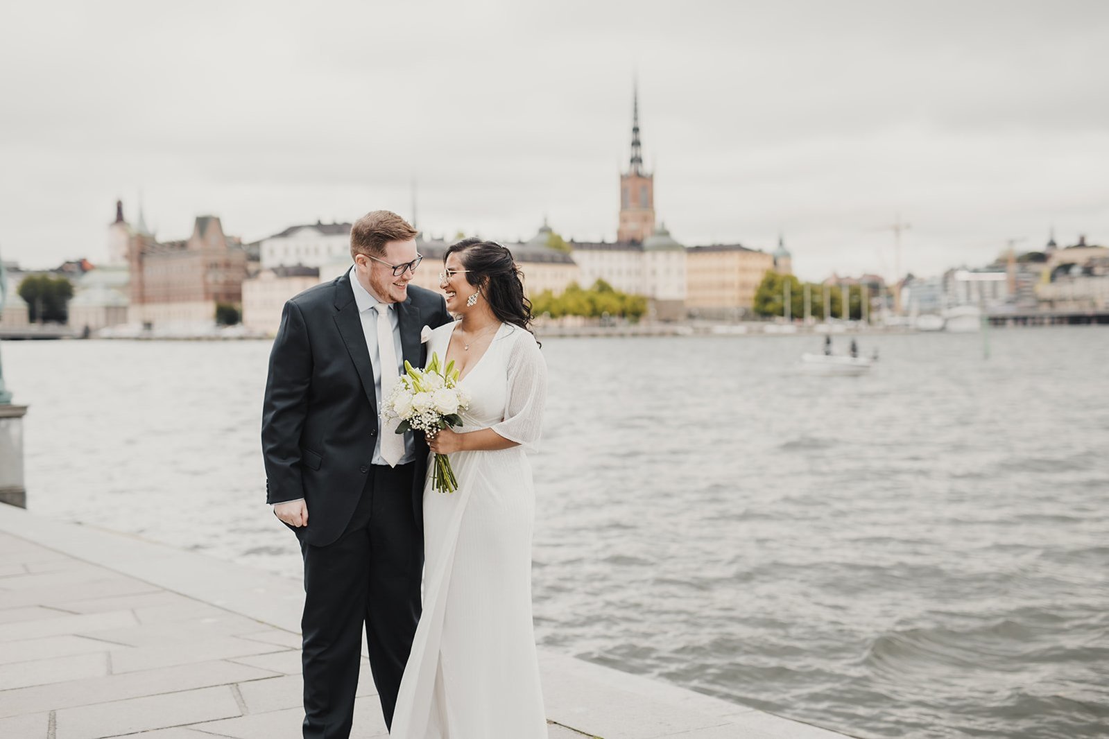Bröllopspar tätt tillammans utanför Stockholms stadshus. Han i blå kostym och hon i vit klänning och brudbukett i handen. I bakgrunden vattnet och ön Riddarholmen.