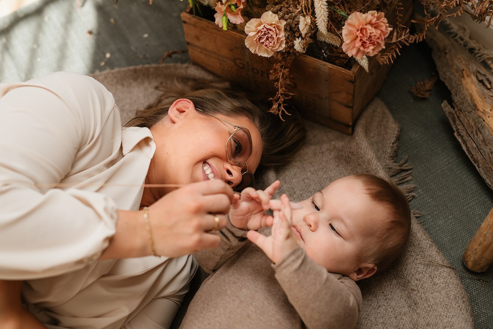Mamma med. beige klänning och. glasögon ligger på en beige filt med sin bebis. Han tittar på växt. hon håller i. Bakom dem nejlikor i en trälåda.