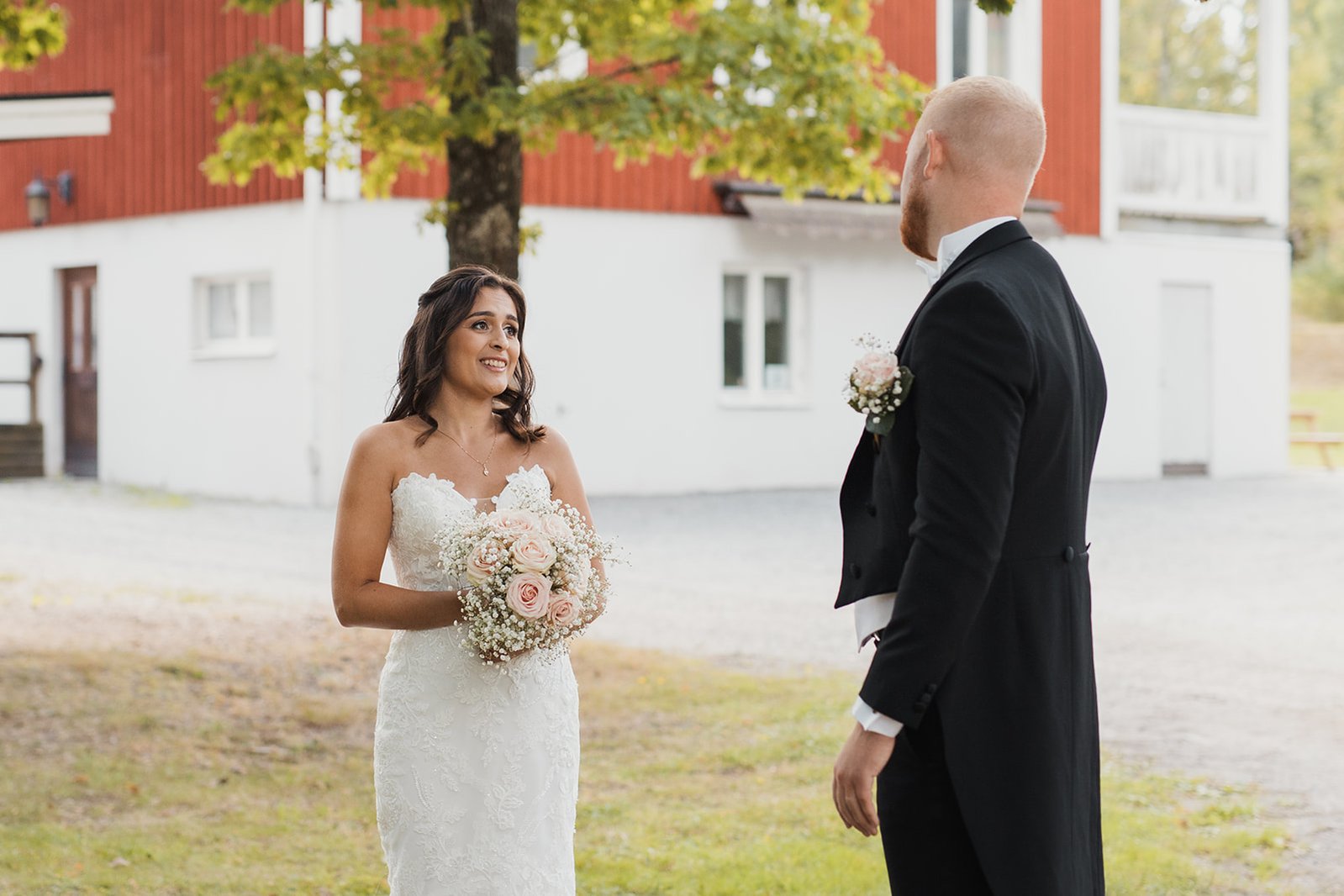 Kvinna i vit ärmlös klänning och brudbukett i handen står och tittar på en man i svart frack utomhus om sommaren. I bakgrunden  hus i falurött med vit puts i grunden.