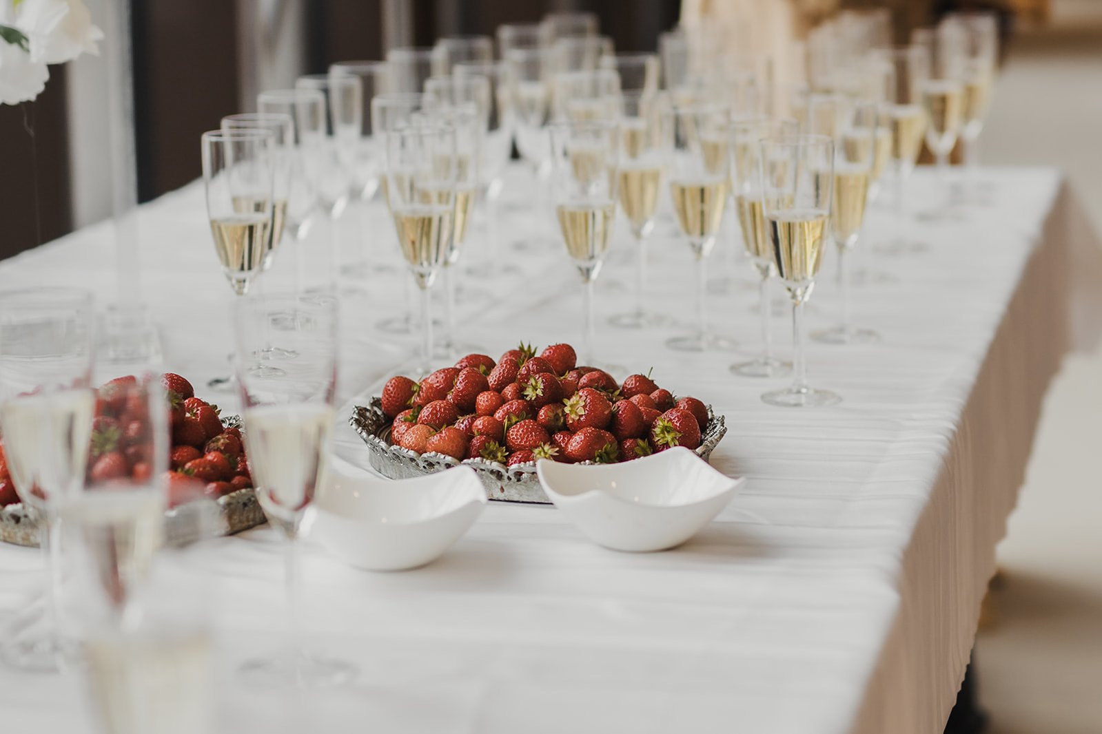 Bord med vit linne duk och massor med champagne glas med bubbel i, 2 silver fat med färska jordgubbar och 2 vita porslin skålar bredvid.