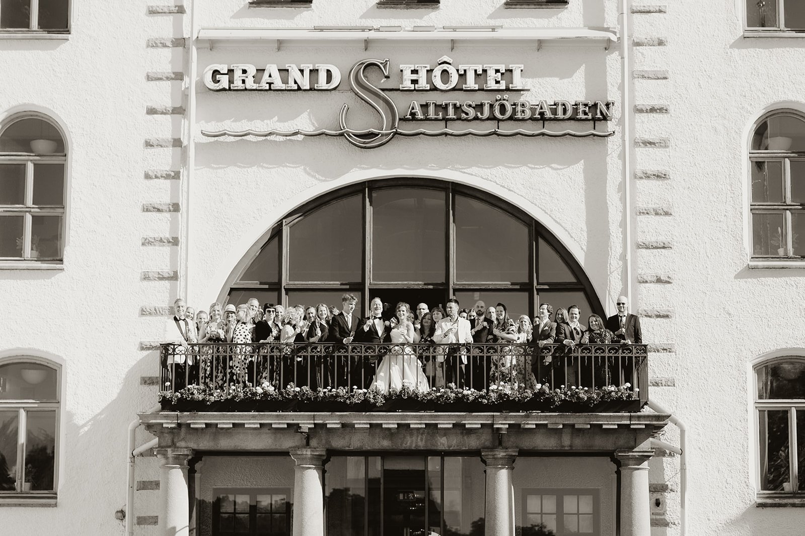  En balkong fullt med gäster och brudpar som skålar, på byggnad en med vi puts och balkongfönster i rund form. Under betongen 4 vita pelare, överst på fasaden skylt med texten "Grand hotel Saltsjöbaden"