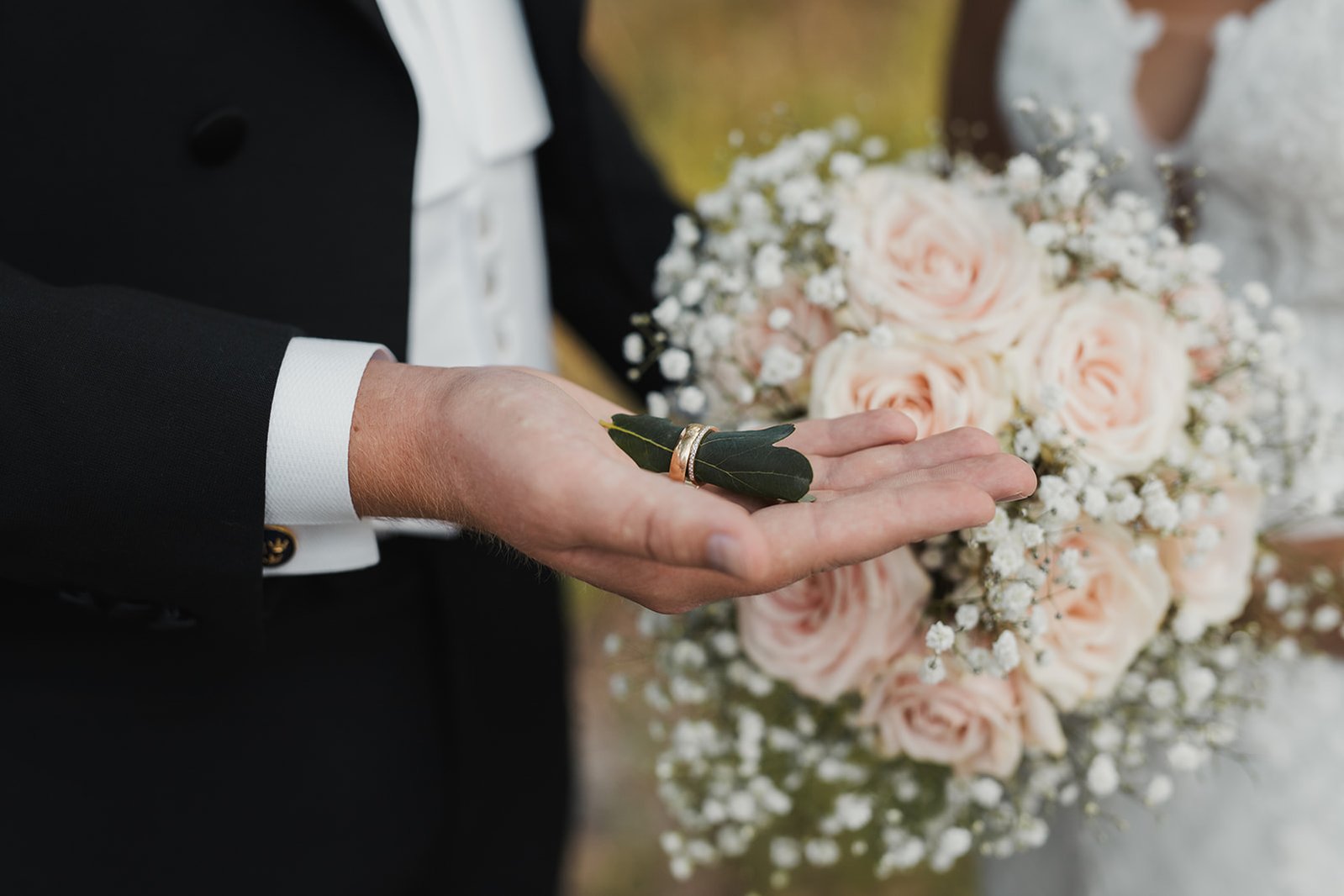 En man hand med handflatan uppåt håller 2 vigselringar trädda i ett grönt löv. I bakgrunden står bruden med brudbukett med rosa blommor och vit brudslöja.