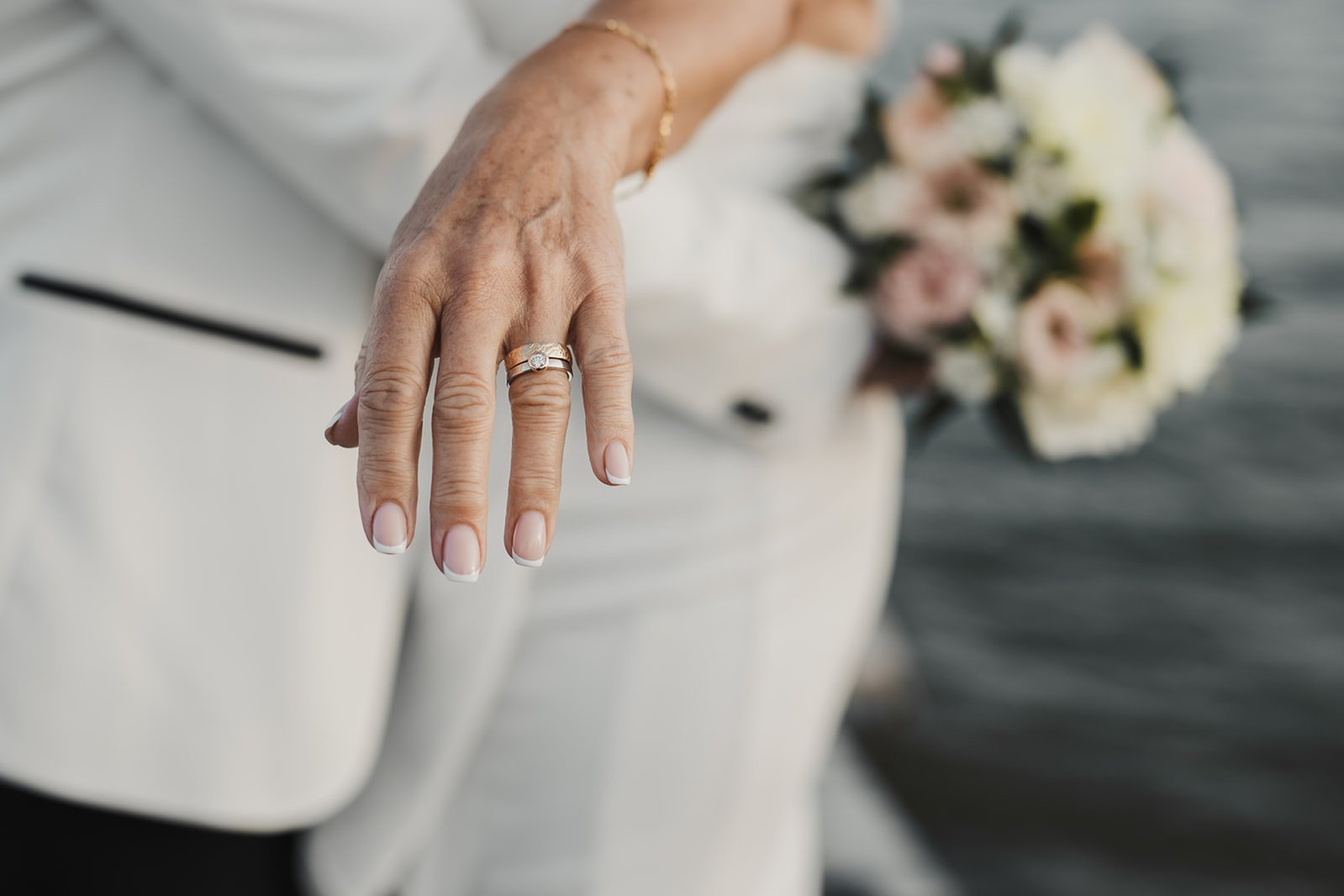En kvinna sträcker ut handen med sin vigselring medan hon kramar om sin man, båda klädda i vitt och med blommor bakom ryggen.