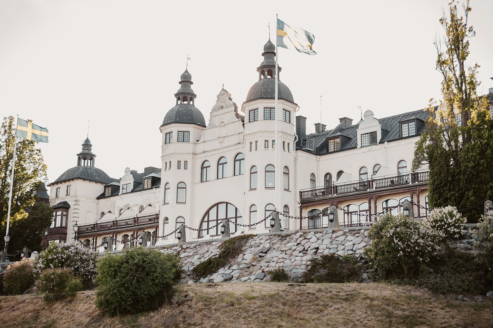 Vit slottsliknande byggnad med 3 torn och svart tak och flera våningsplan och balkong. Omgärdas av gräsmatta, buskar,  2 flaggstänger med svenska flaggan.