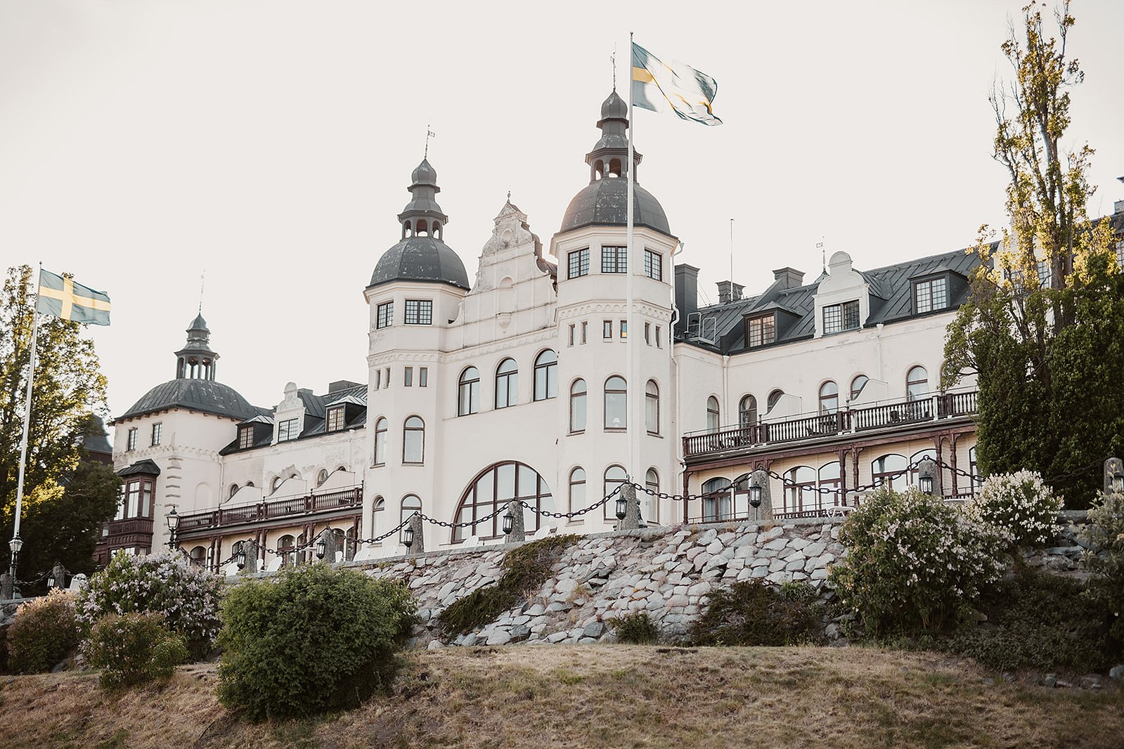 Vit slottsliknande byggnad med 3 torn och svart tak och flera våningsplan och balkong. Omgärdas av gräsmatta, buskar,  2 flaggstänger med svenska flaggan.