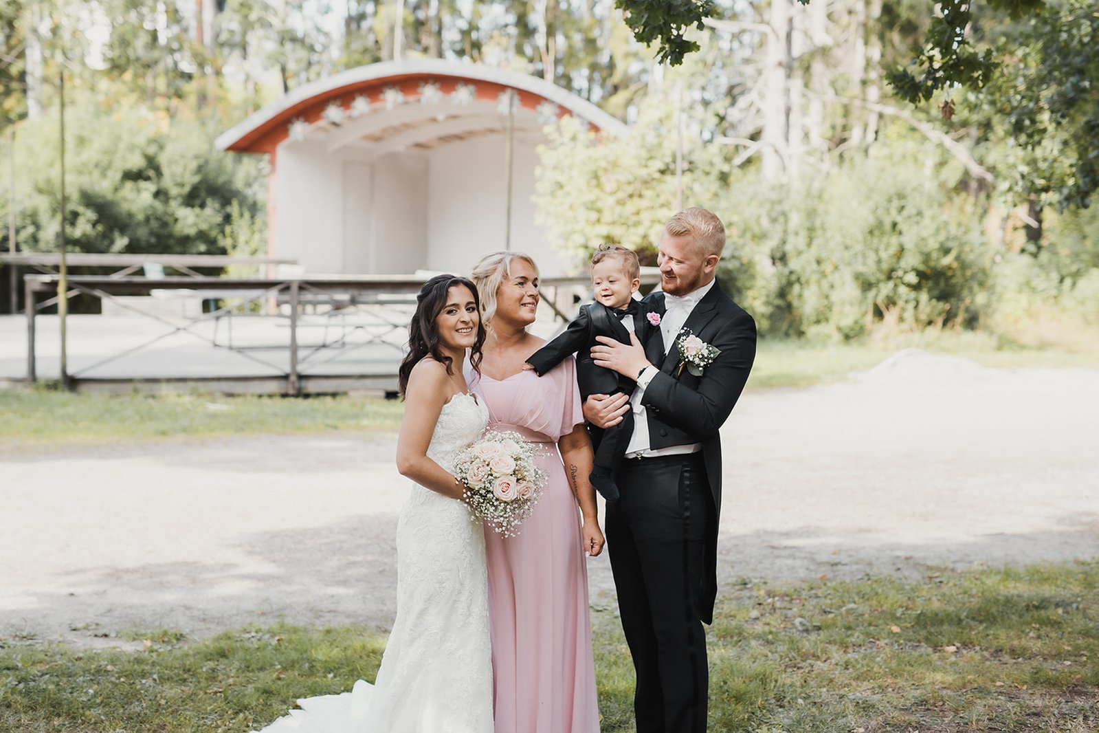 Kvinna i vit lång klänning med blommor i handen, bredvid hennes mor i rosa klänning. Man i familjen med svart frack håller sin son i famnen. Dem står tillsamman utomhus framför en dansbana.