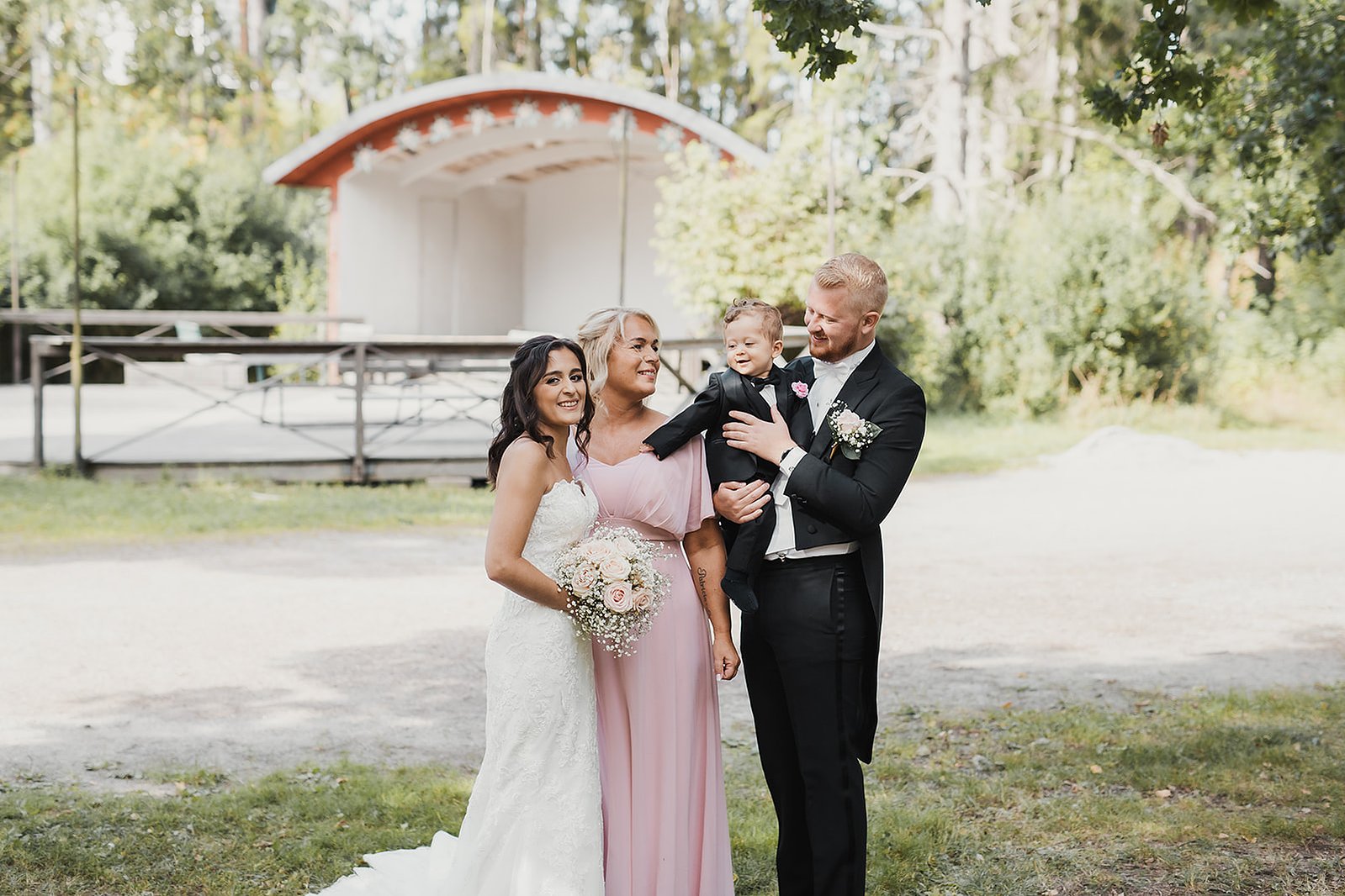 Kvinna i vit lång klänning med blommor i handen, bredvid hennes mor i rosa klänning. Man i familjen med svart frack håller sin son i famnen. Dem står tillsamman utomhus framför en dansbana.