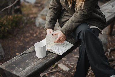 Kvinna i grön jacka och svarta byxor sitter på en träbänk utomhus, bläddrar i skrivbok med vita blad med anteckningar och har en vit kaffemugg på bänken.