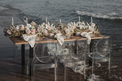 Dukat bord på stranden med vågorna under dem genomsskinliga stolarna. Bordet med träskiva är festligt dukat för 3 på var sida med blommor, ljusstakar, tallrikar,bestick och glas.