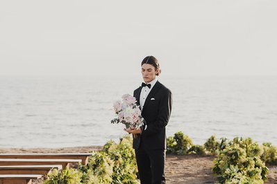 Man i svart kostym, svart flyga och vit skjorta väntar på sin brud med brudbuketten i handen. Han står på en sandstrand med träbänkar och grönska bakom havet.