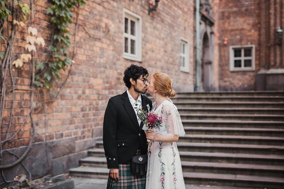 Man i mörkt lockigt hår och glasögon kysser kvinna med rött hår uppsatt i en knut. Hon är klädd i vit klänning med blommor på och håller brudbukett i handen. Bakom dem röd tegelbyggnad med en trappa.