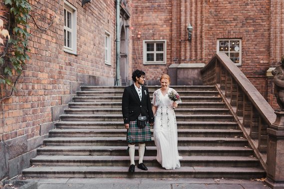 Man med glasögon, lockigt hår och kilt går ner för en trappa hand i hand med en kvinna i vit klänning, rött uppsatt hår och bukett i handen. Bakom dem röd tegelbyggnad med vita fönster.
