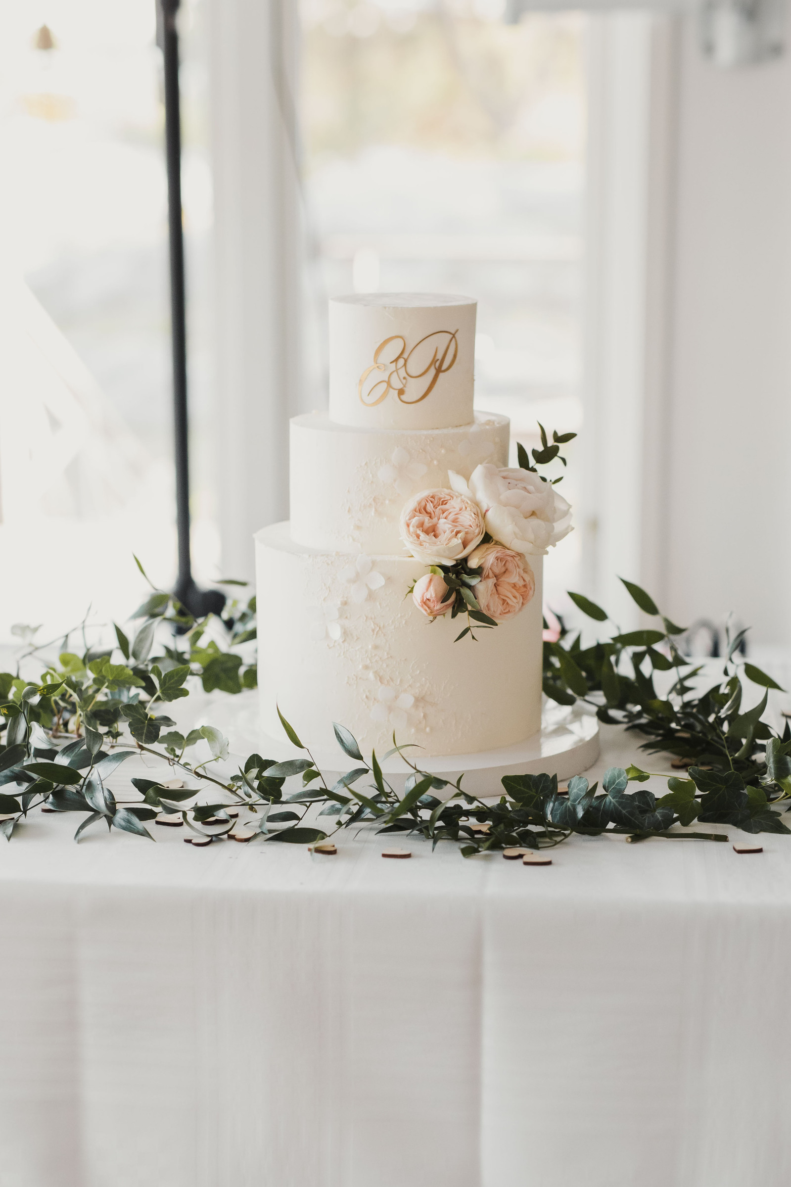 Bröllopstårta i 3 våningar i vitt, dekorerad med rosa blommor och brudparets initialer E och P. Tårtan är placerad på bord med vit duk  i festlokalen på restaurang Solbrännan. Bakom skymtar fönster stativ till högtalare.