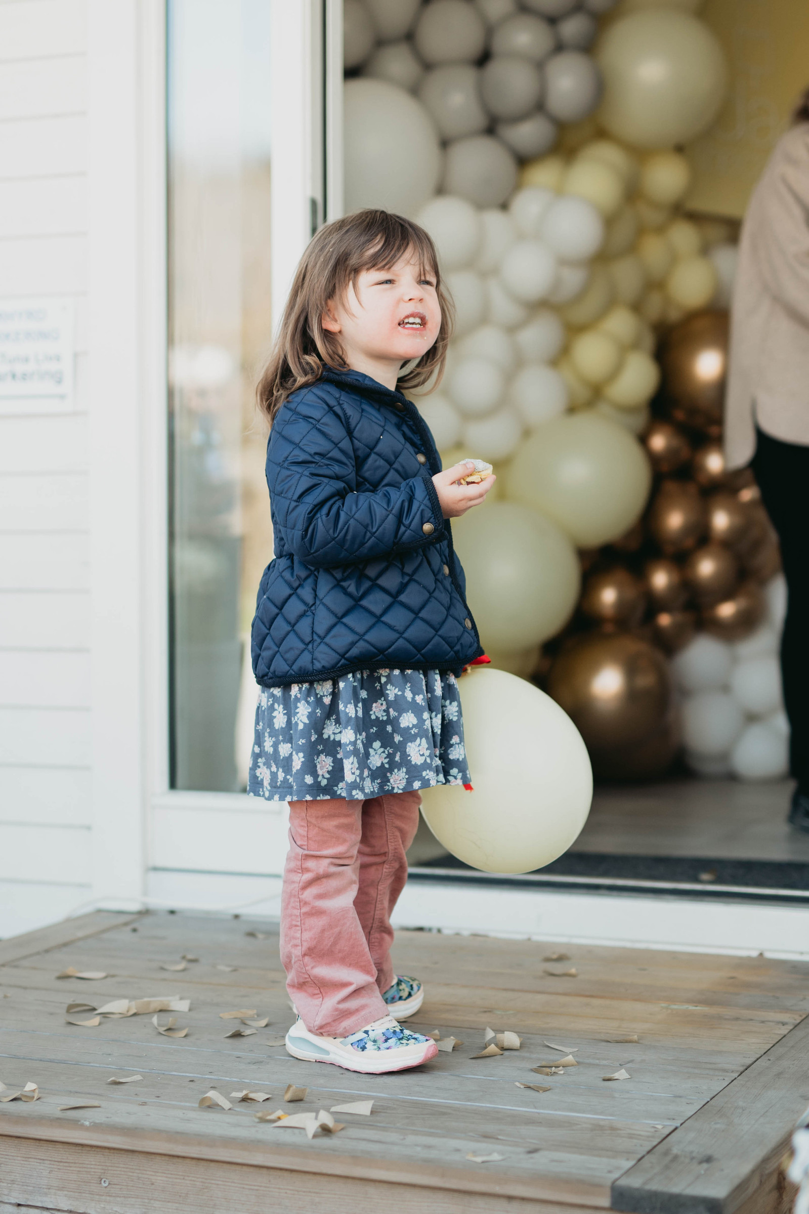 Flicka ii blå jacka och rosa byxor håller i en gul ballong och en cupcake i handen stående i en trätrappa. Bakom ingång till studio -  en ballongvägg.