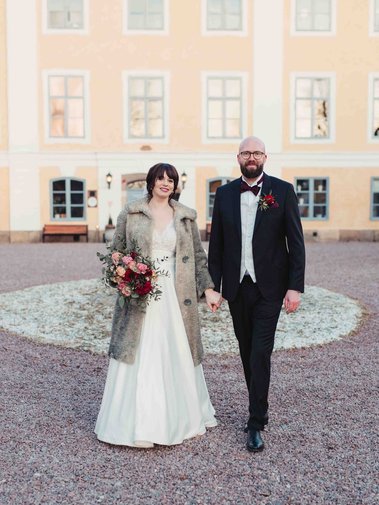Brudpar framför en herrgård med gula fasader. Kvinna i lång vit klänning och beige vinterjacka, håller en brudbukett i handen. Han i mörk kostym och fluga håller i hennes hand medan dem pratar gåendes på grusgången.