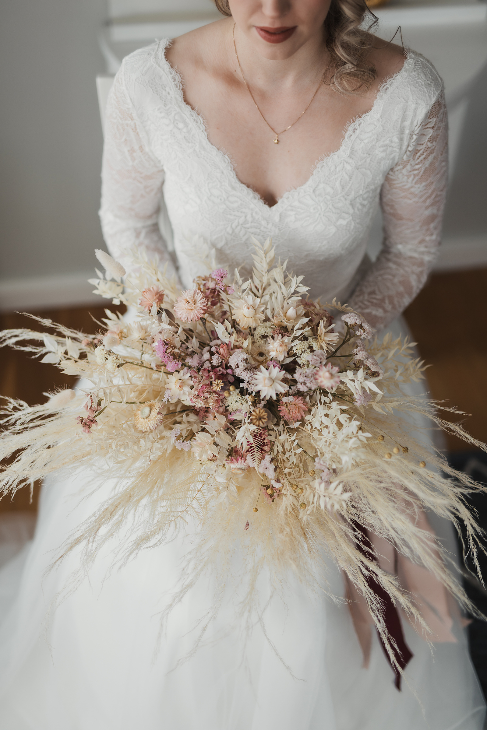 Bukett med torkade blommor i beige och rosa färg och siden band- hålls av en sittande kvinna i vit lång klänning.