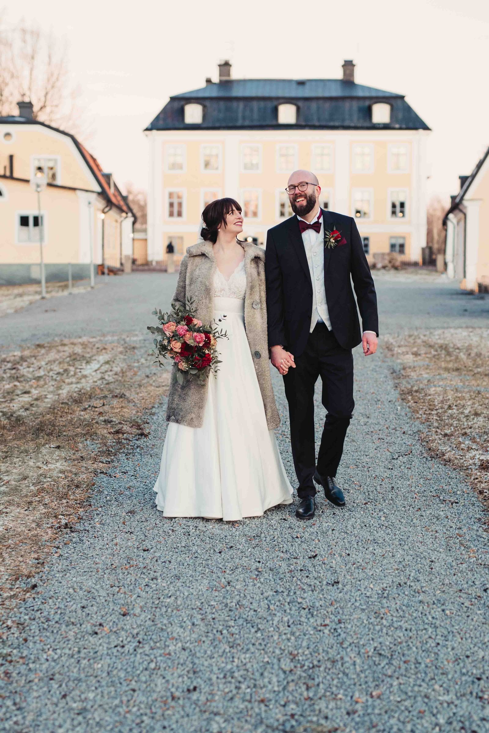 Bröllopspar går hand i hand på en grusgång framför en herrgård. Han i mörk kostym och glasögon, hon i lång vit klänning och vinter rock - håller sin brudbukett i andra handen.