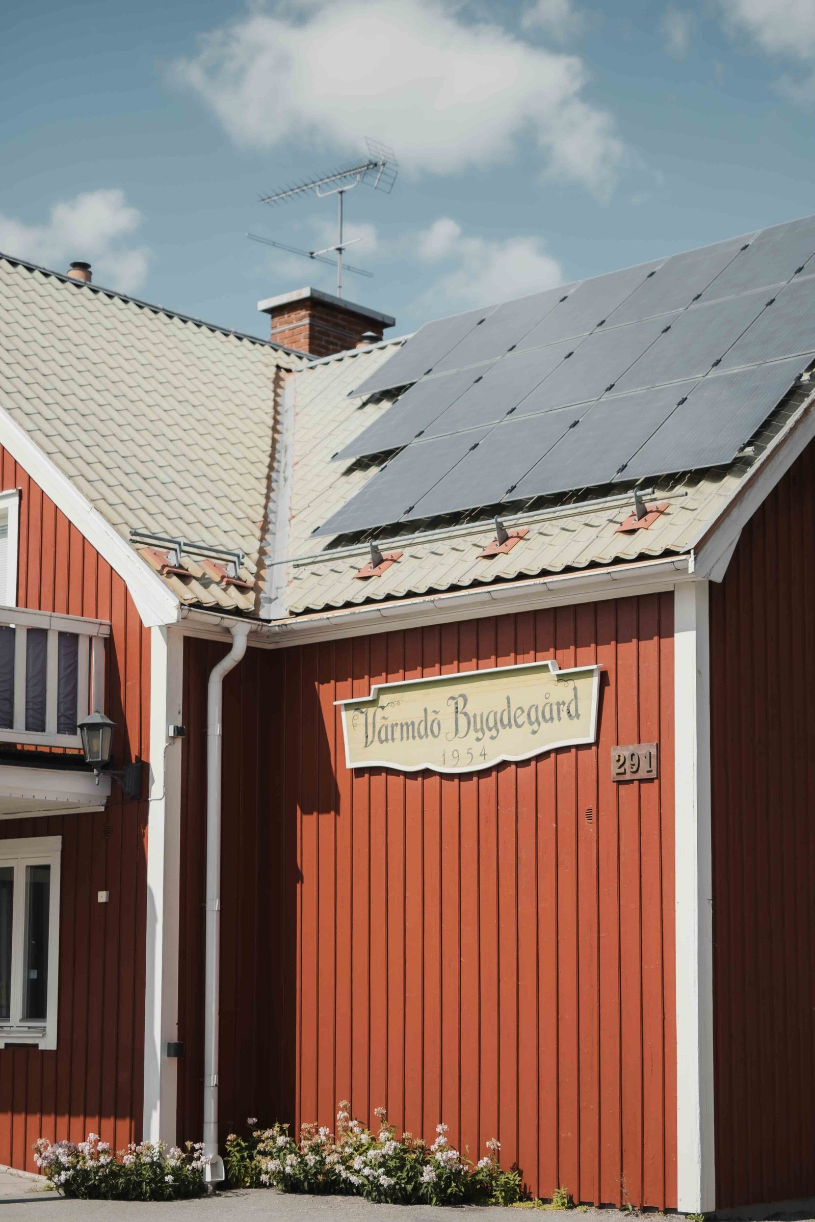 Trä byggnad i falurött och vitmålad knut, tak med solpaneler och antenn.  Stupränna och skylt med texten "Värmdö bygdegård. 1954". Blommor planterade intill husväggen.