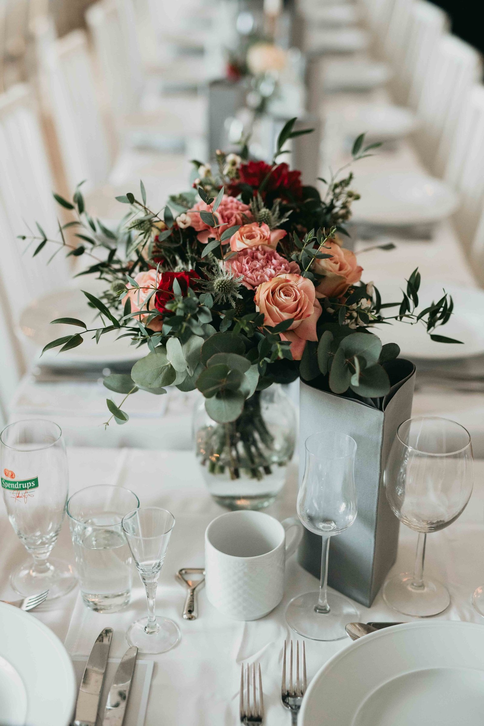 Dukat långbord för bröllopsfest med blommor från tärnornas bukett i festlokalen på Schenströmska herrgård. Bord med vit duk dukat med tallrikar,bestick, glas och koppar på dukade bordet.
