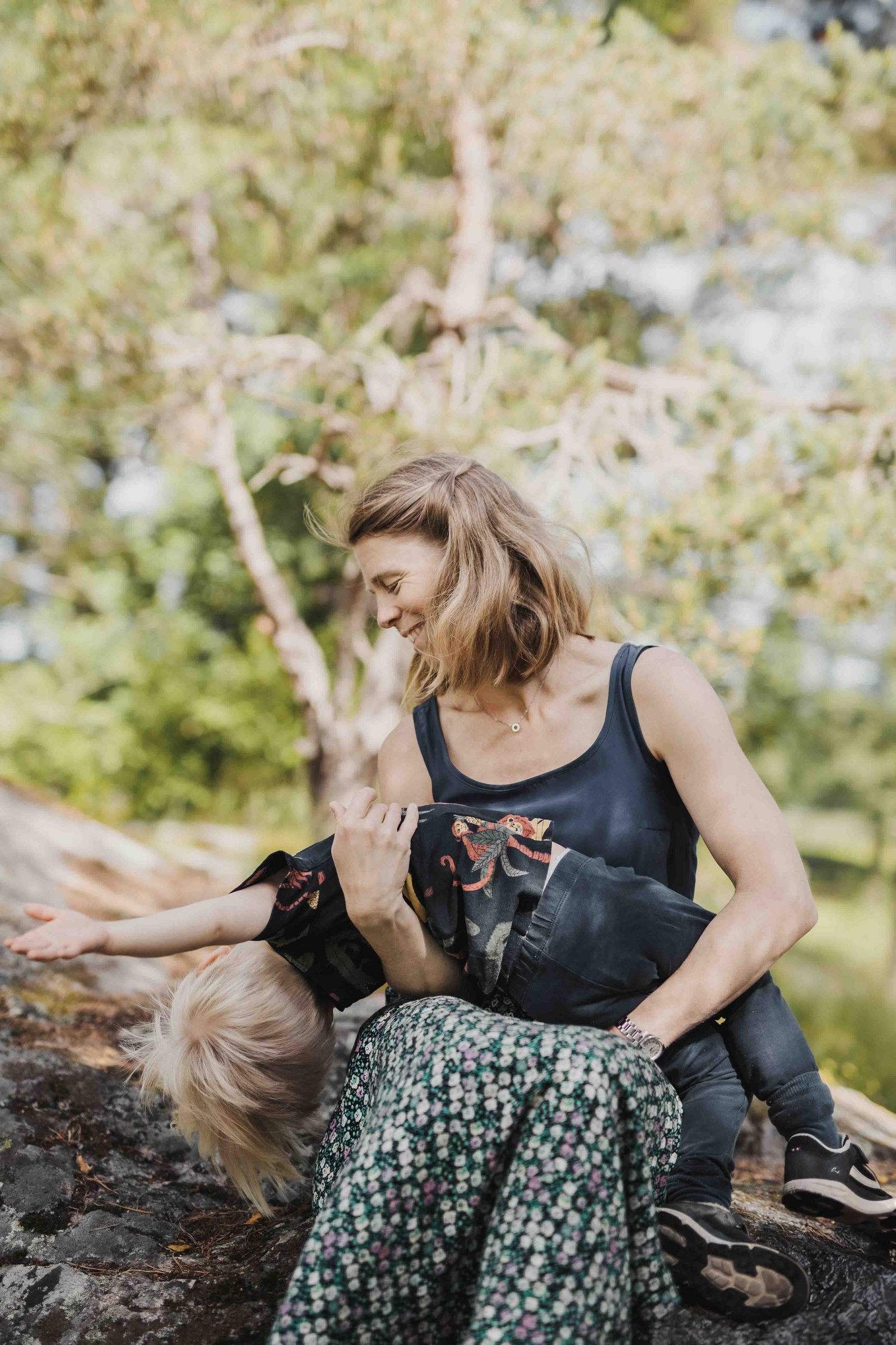 Kvinna sitter med sitt barn i famnen utomhus sittandes vid en sten i skogen. Hon är klädd i mörkblå topp och mönstrad kjol. Pojke med ljust hår. blå byxor och blå t-shirt med djurmotiv.
