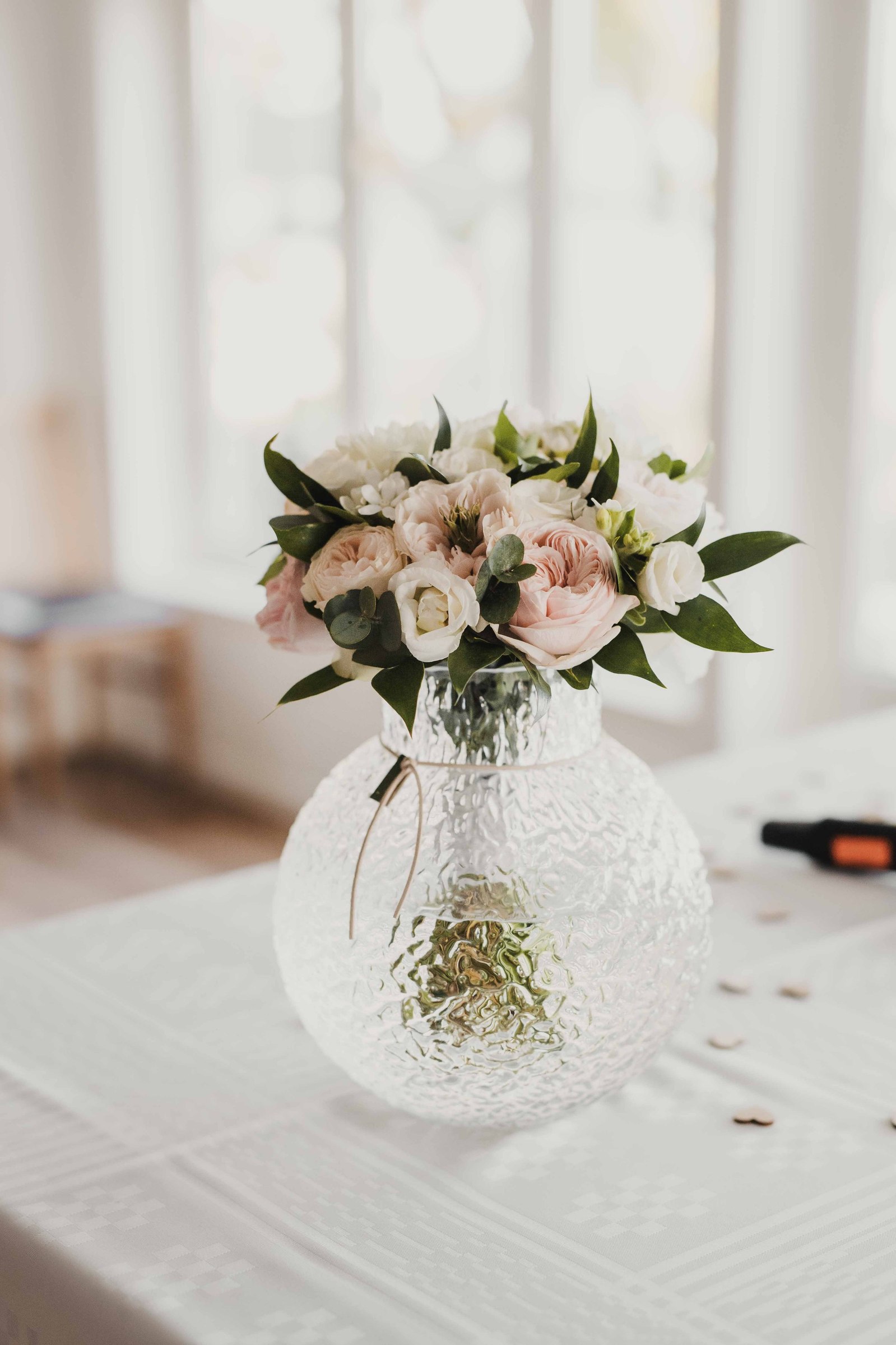 Brudbukett med rosa rosor, vita blommor och gröna blad i rund glasvas placerad på dukat bord med vit duk, i festlokalen på restaurang, i bakgrunden stora fönster med mycket ljusinsläpp.