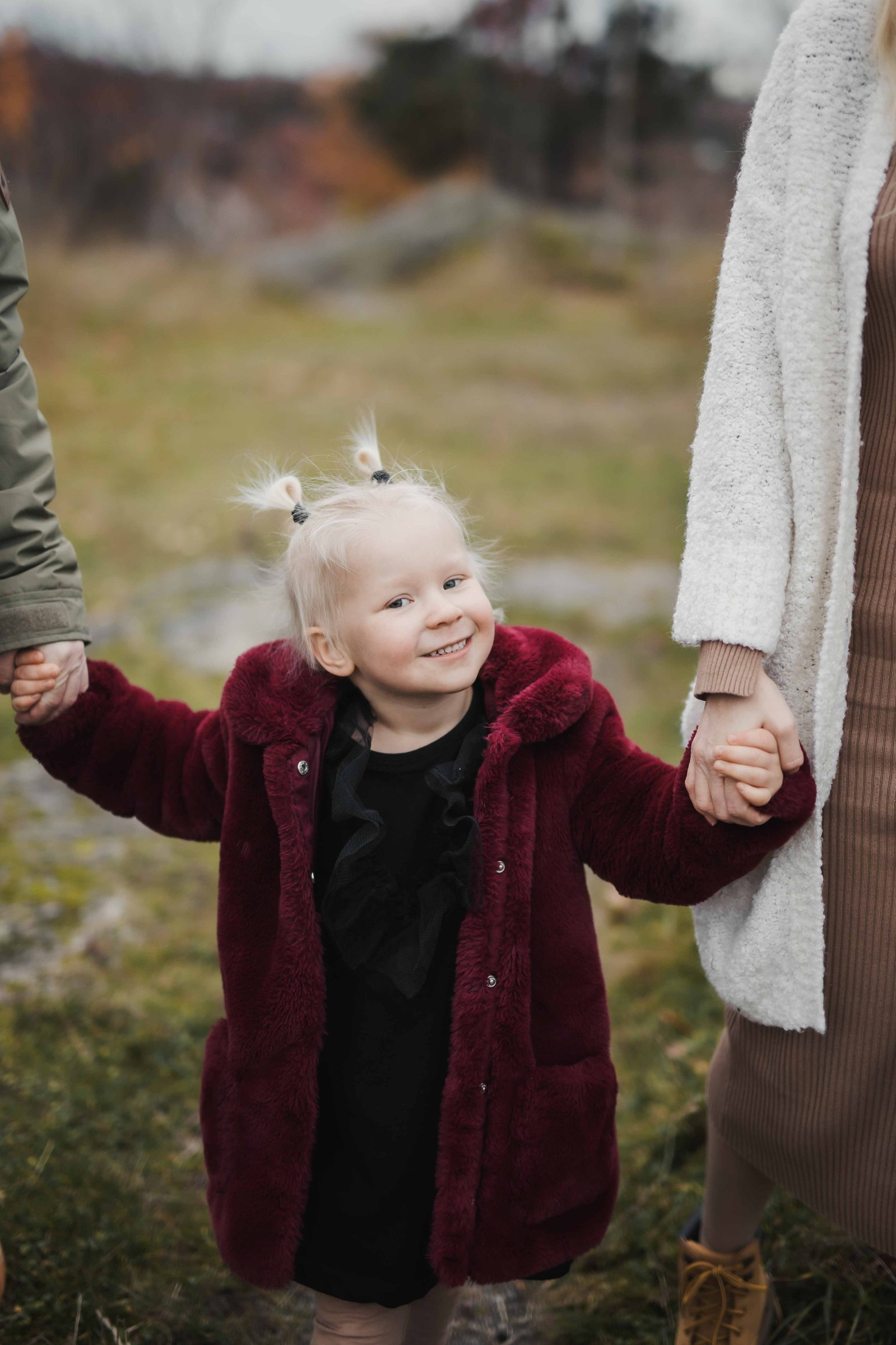Flicka klädd i vinröd rock och svart klänning med 2 blonda tofsar i håret, håller sina föräldrar i handen. på promenad utomhus.