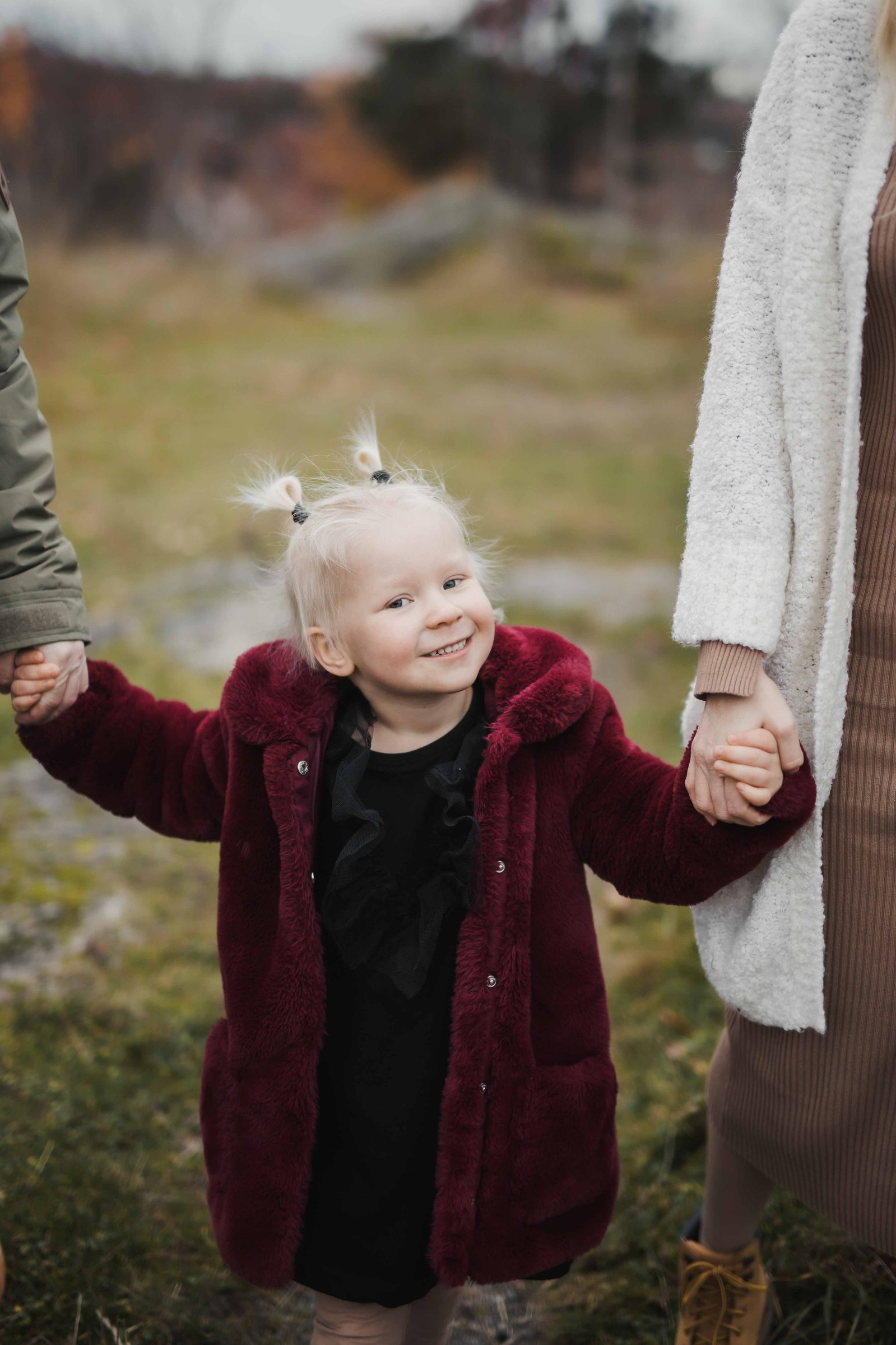 Flicka klädd i vinröd rock, svart klänning , 2 tofsar i det blonda håret håller varsin förälder handen när dem går utomhus på gräsmatta.