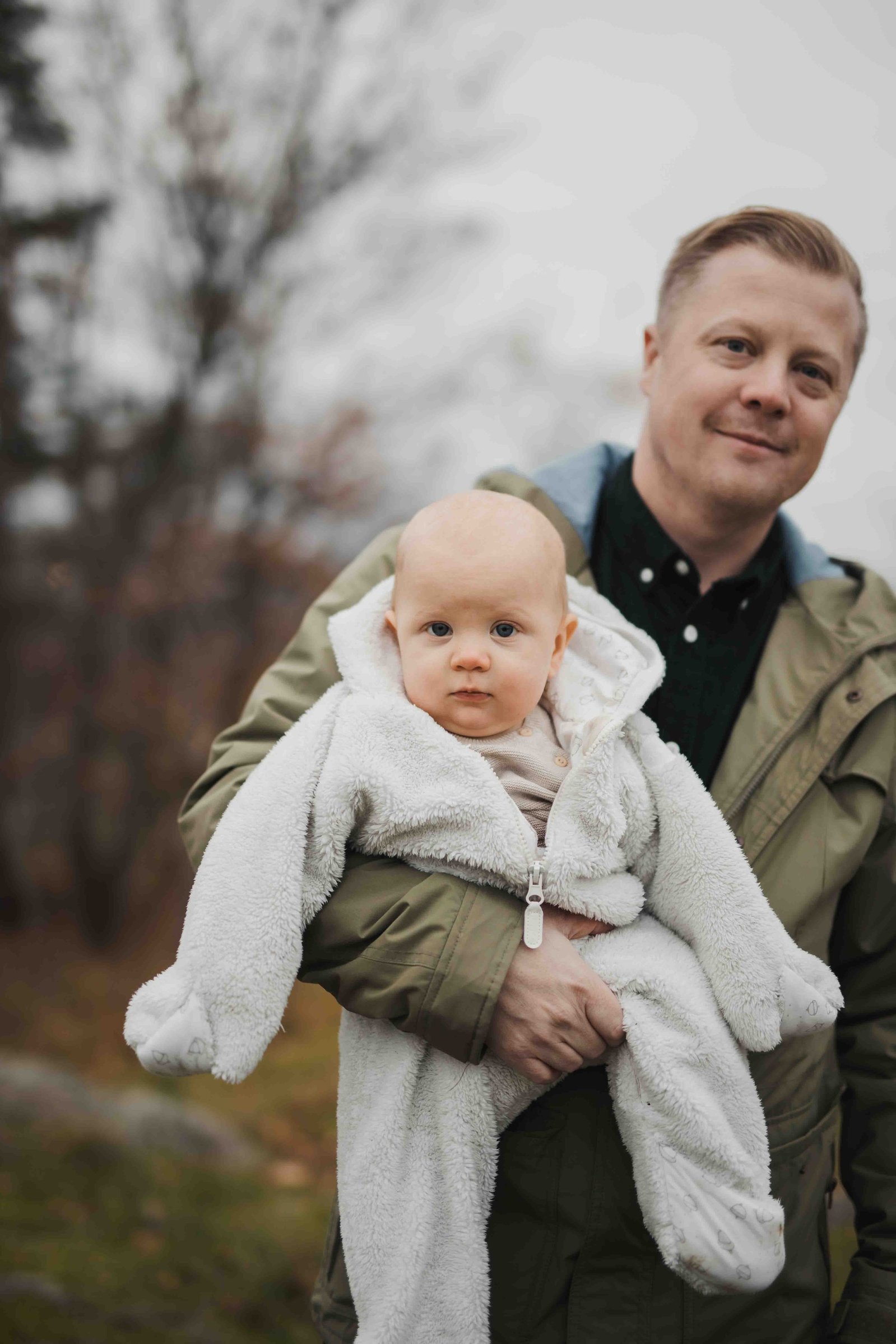 Bebis i vit overall hålls i famnen av sin pappa, pappa i grön jacka och svart skjorta.