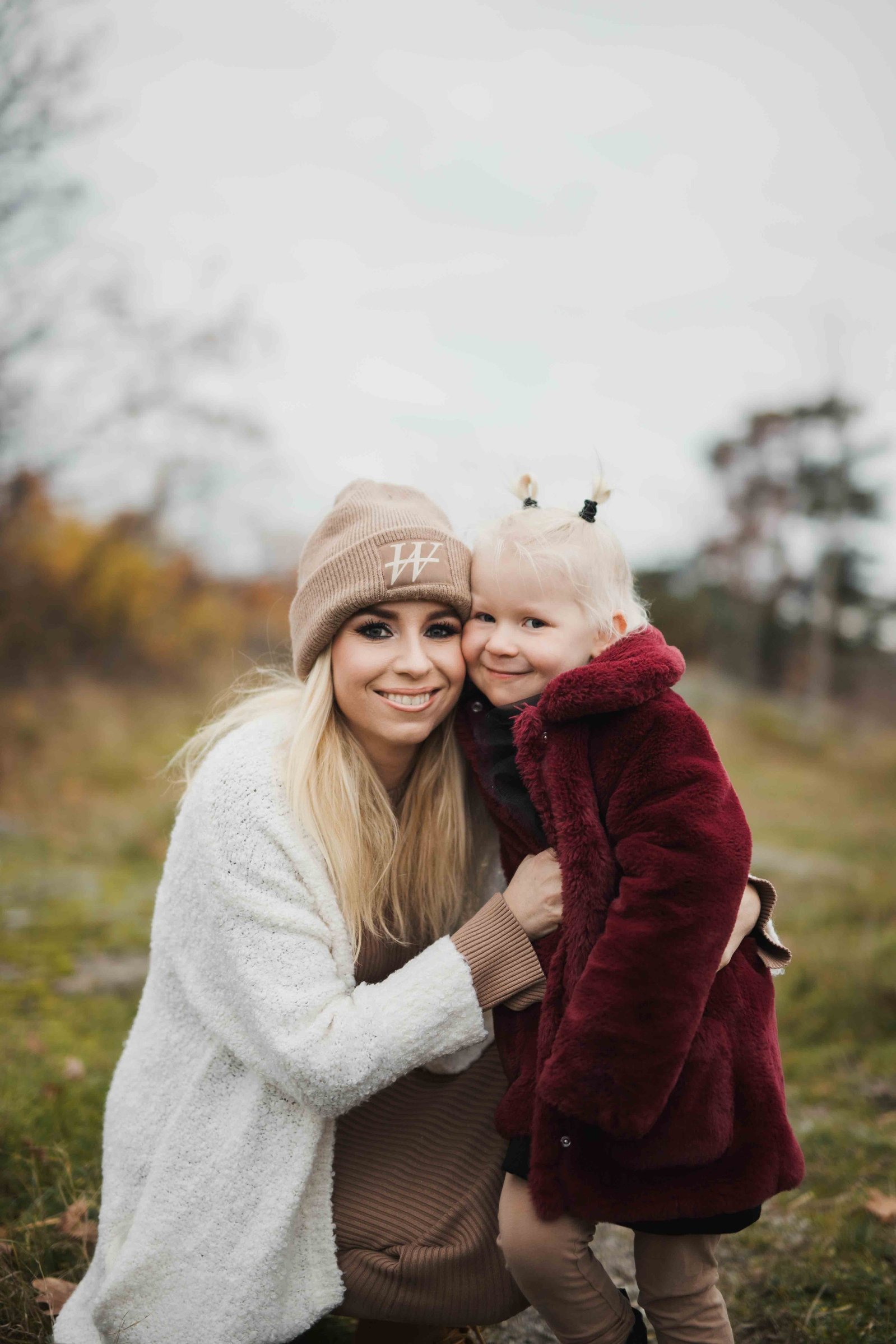 Mamma i vit kofta och beige stickad mössa kramar om sin dotter, klädd i vinröd rock och 2 tofsar i det blonda håret utomhus på hösten.