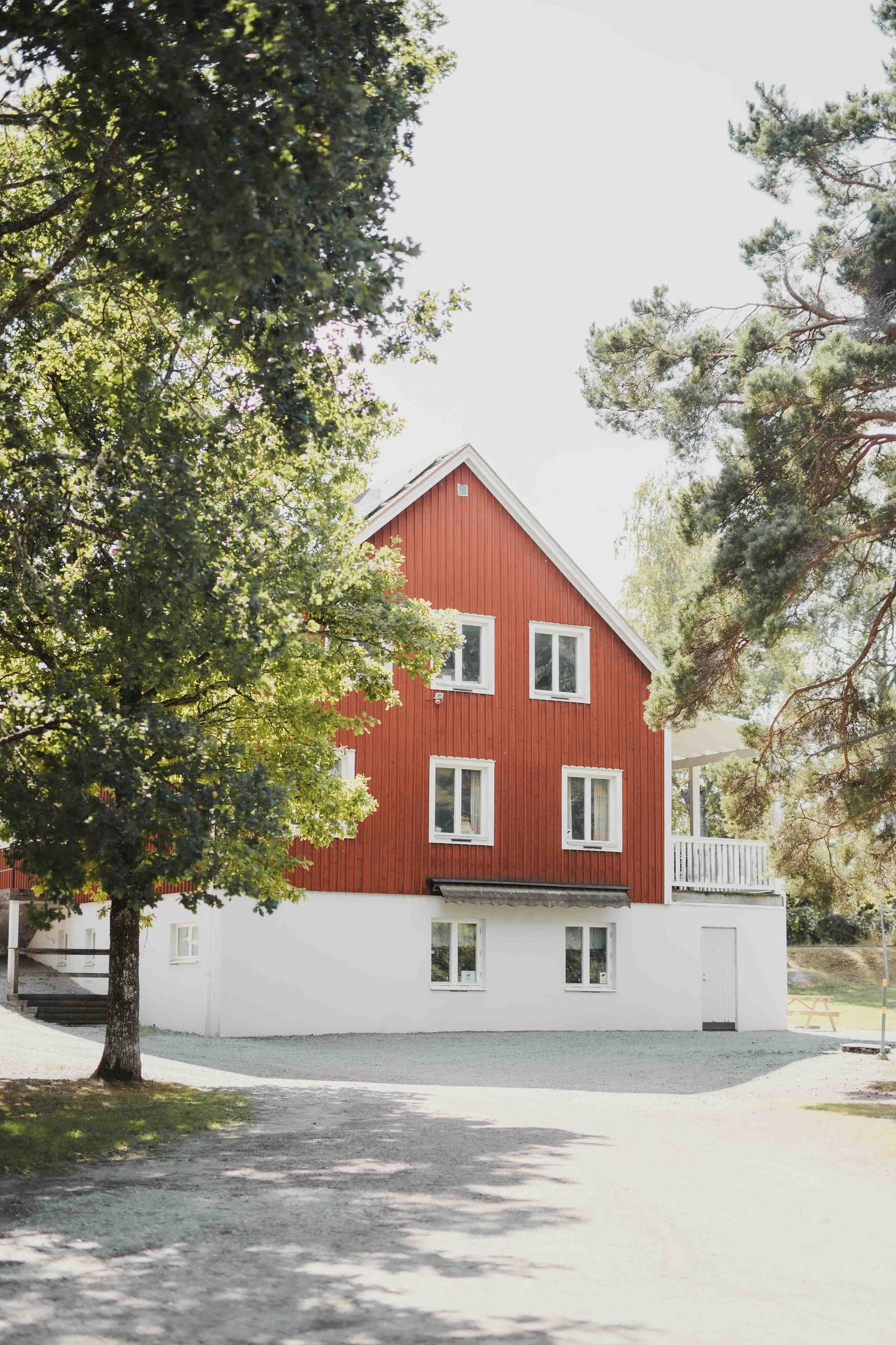 Trä byggnad i två våningar i falurött och vit puts i grunden omgiven av träd i grönska.